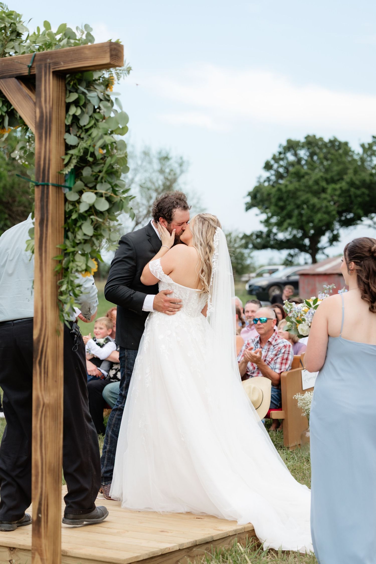 Bride and groom share first kiss under rustic wooden arch at outdoor farm wedding ceremony.