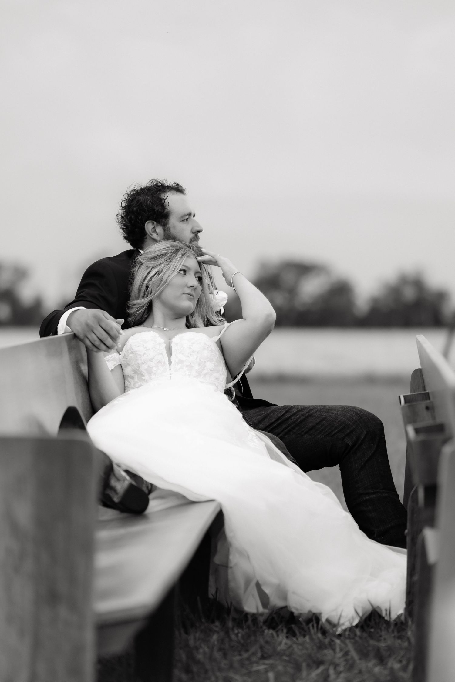 Black and white portrait of bride and groom sitting together on wooden bench at outdoor wedding.