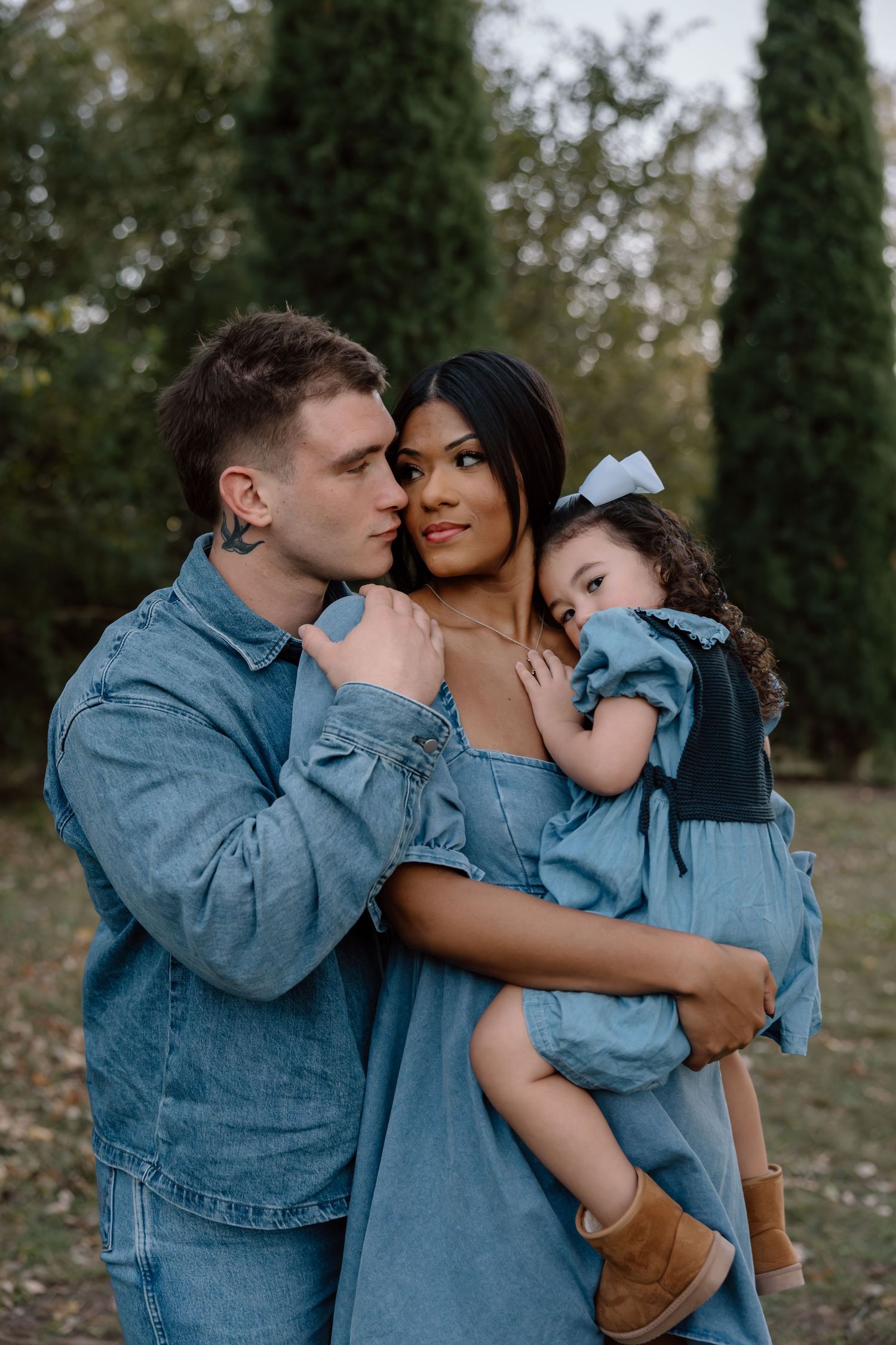 A loving couple in denim outfits embraces while holding their sleepy toddler daughter in a garden setting.