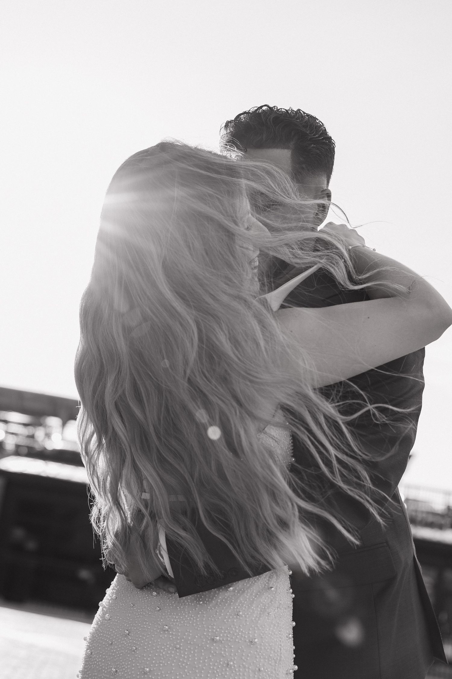 Black and white romantic photo of couple embracing with sun flare and woman's long hair blowing.