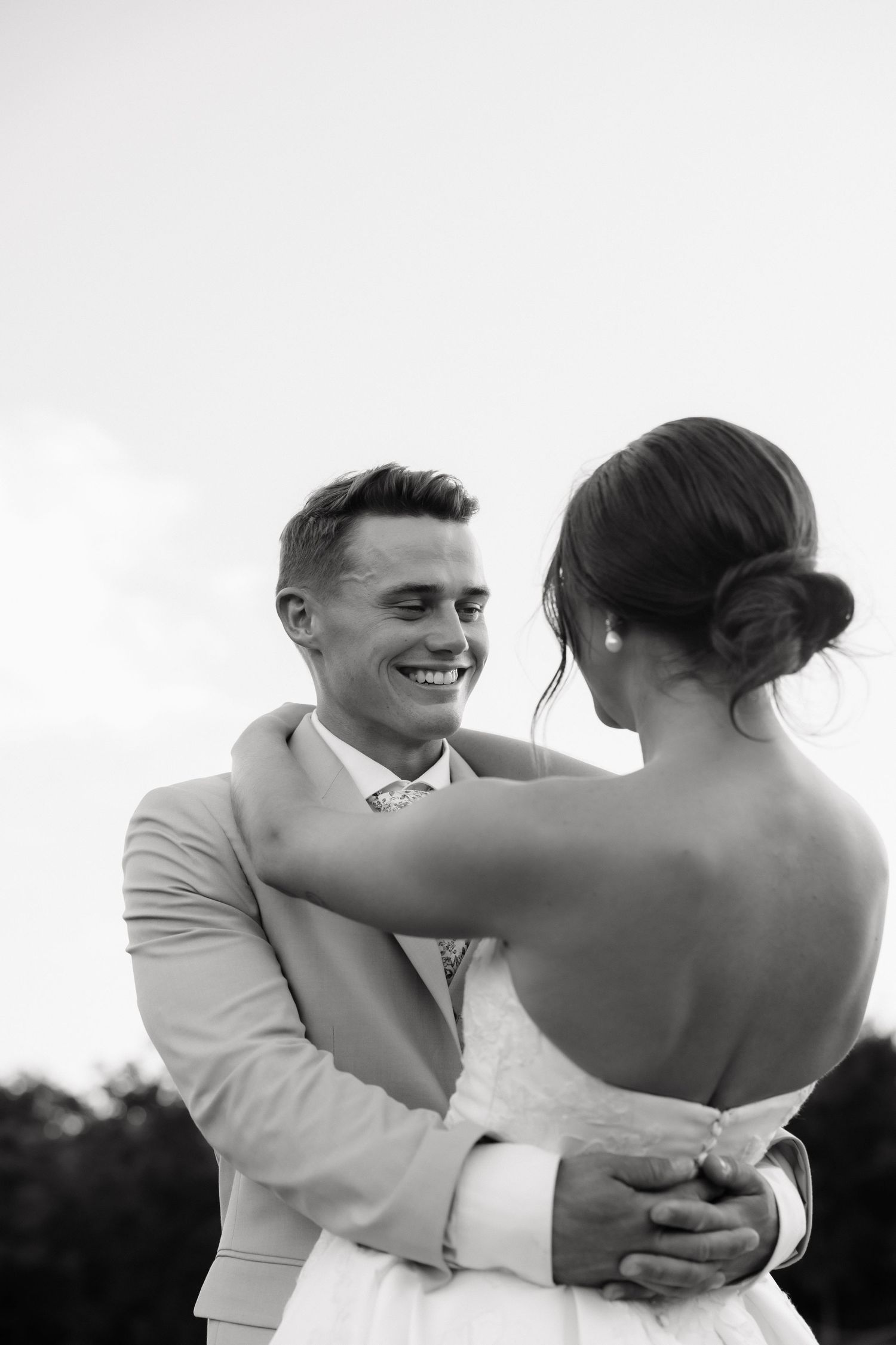 Black and white portrait of smiling groom embracing bride outdoors against a soft sky background.