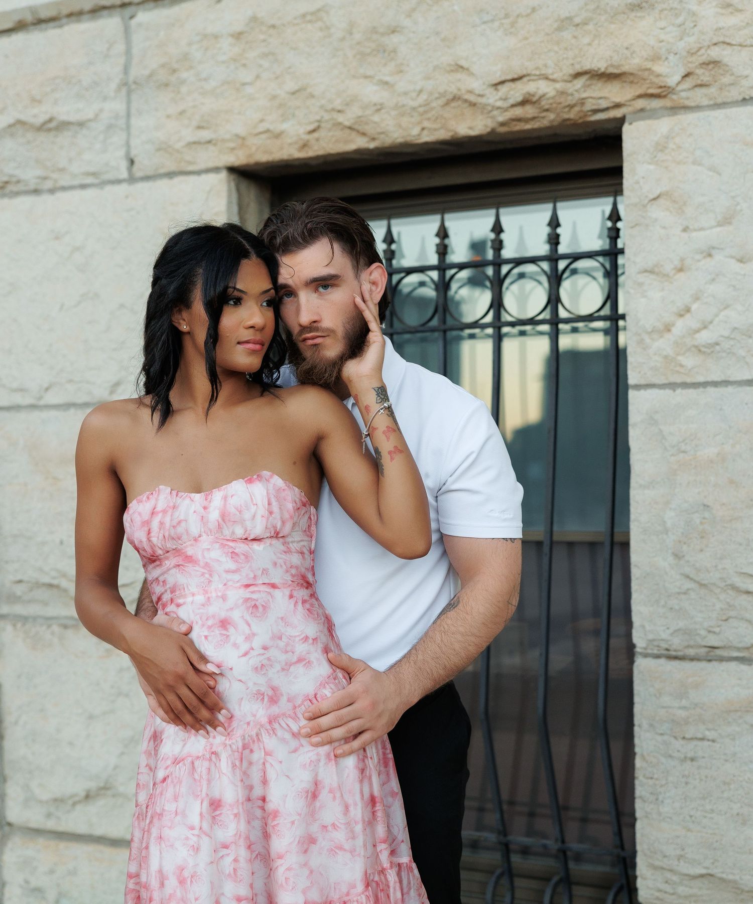 Elegant interracial couple posing by a stone wall, woman in pink floral strapless dress at sunset.
