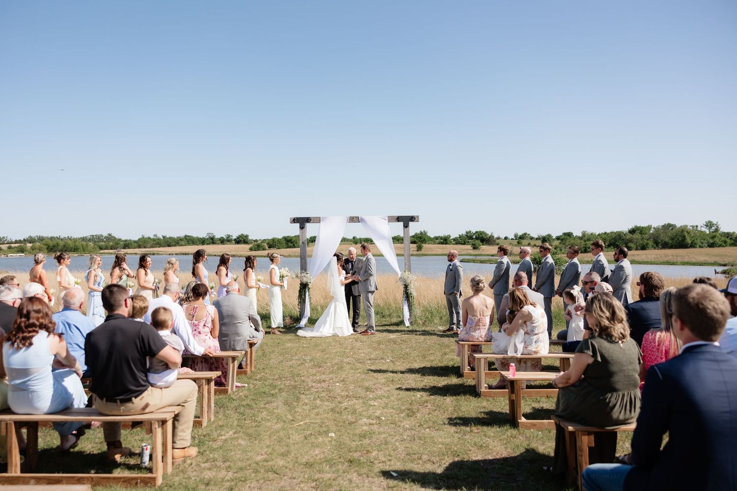 Outdoor wedding ceremony by a lake with guests seated on wooden benches.