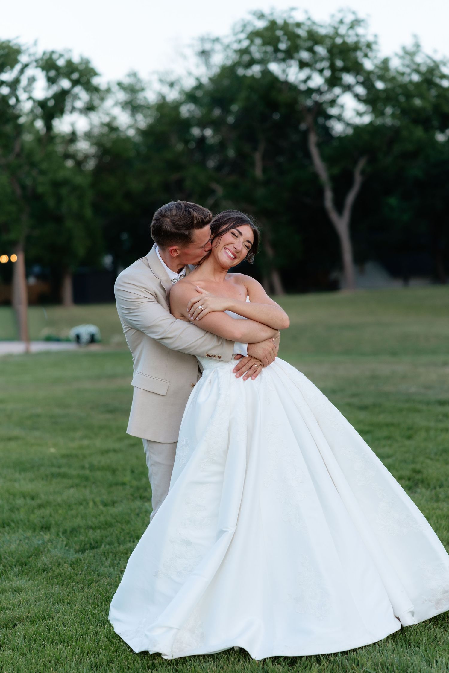 Groom in tan suit embracing bride in full white ball gown on lush green lawn at twilight.