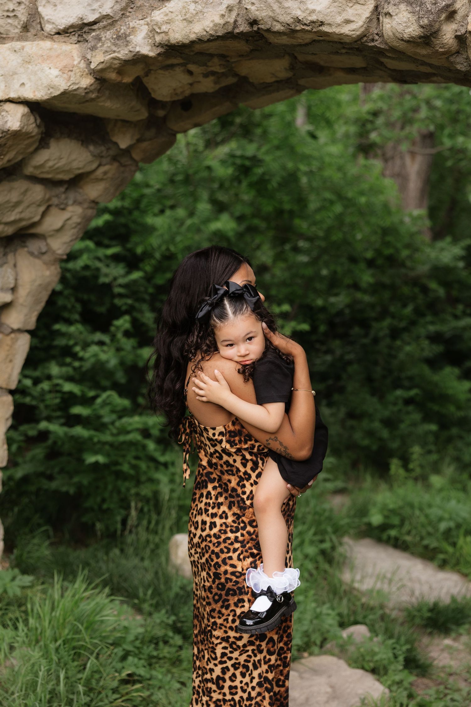 A woman in a leopard print dress holds a young child under a stone arch surrounded by green foliage.