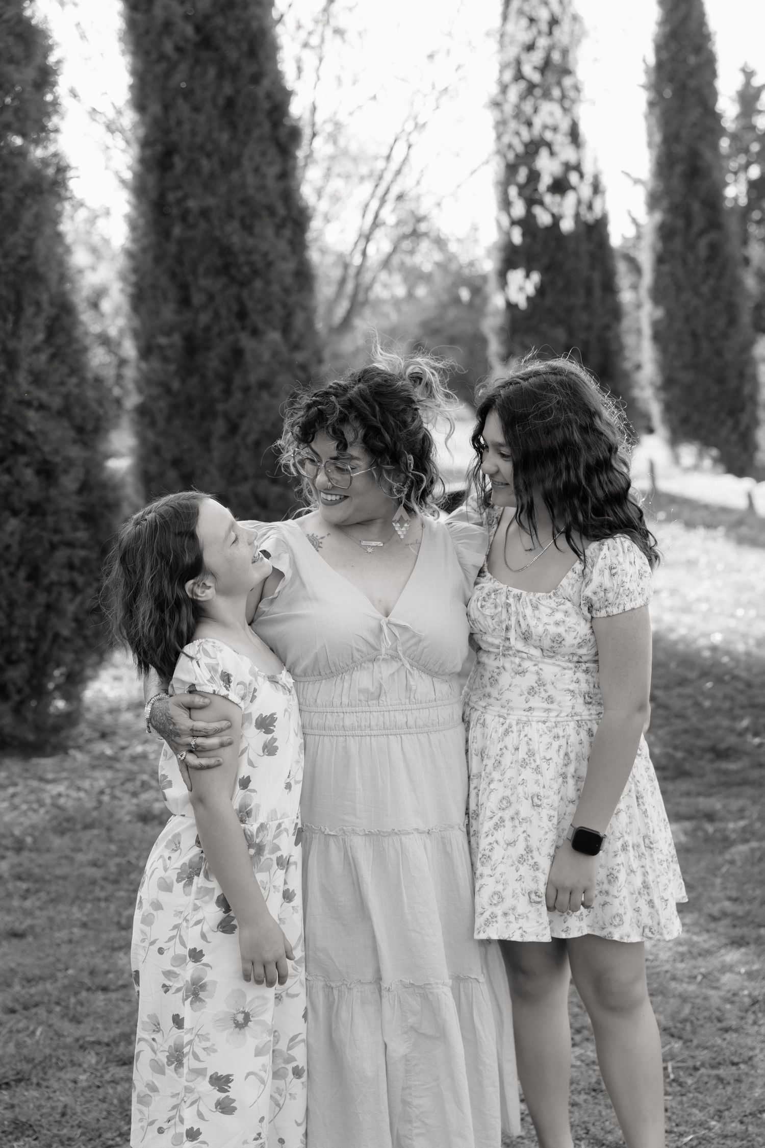 Black and white photo of a woman hugging two girls while laughing together in a garden with tall trees.