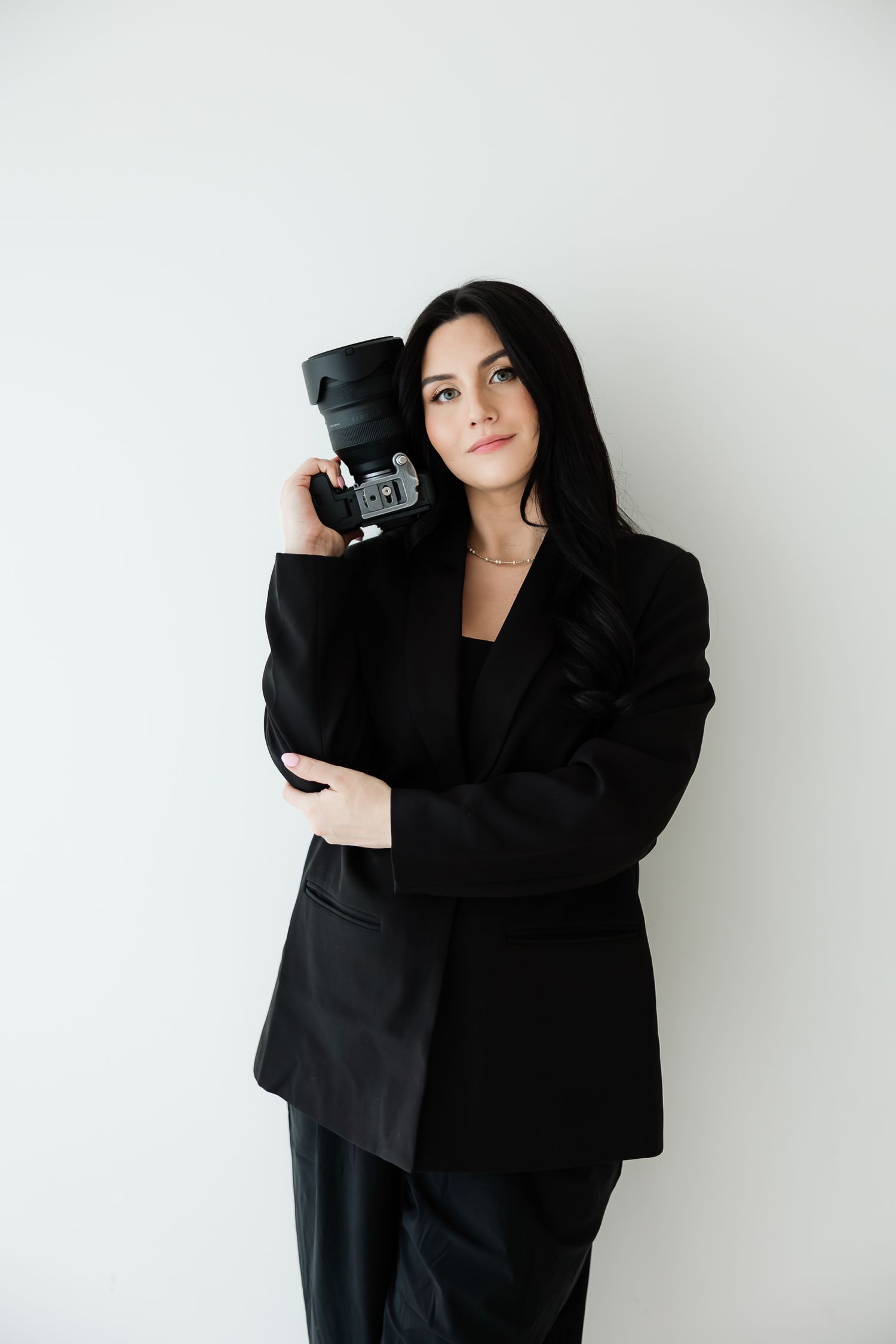 Professional female photographer in black suit holding camera against white background.