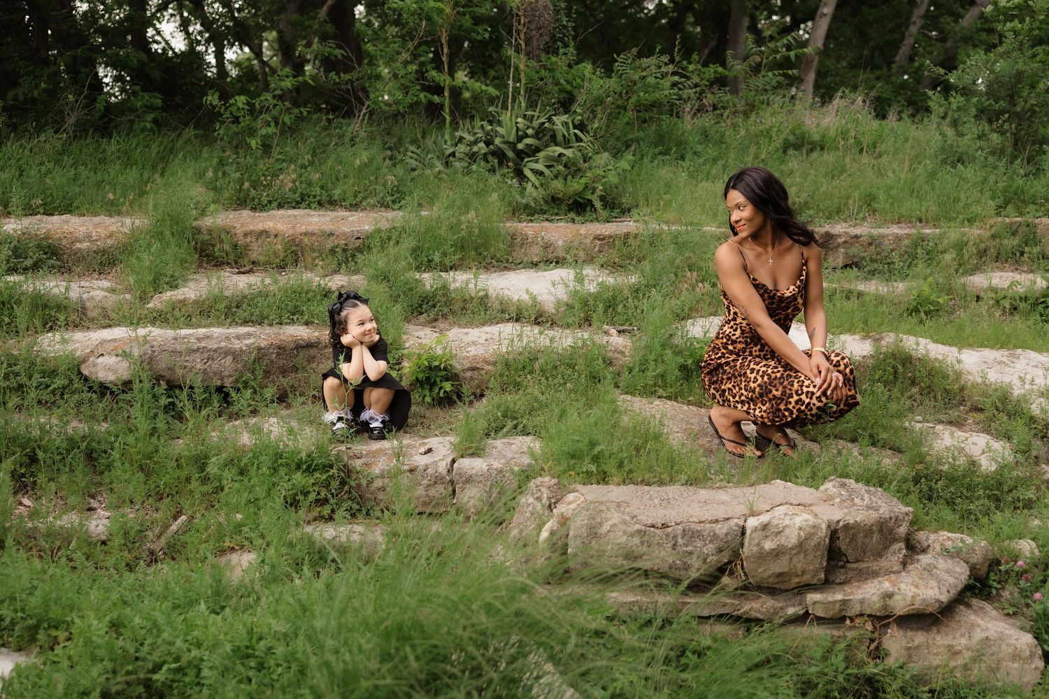 A woman in a leopard print dress crouches near a young girl sitting on stone steps in a lush green park.