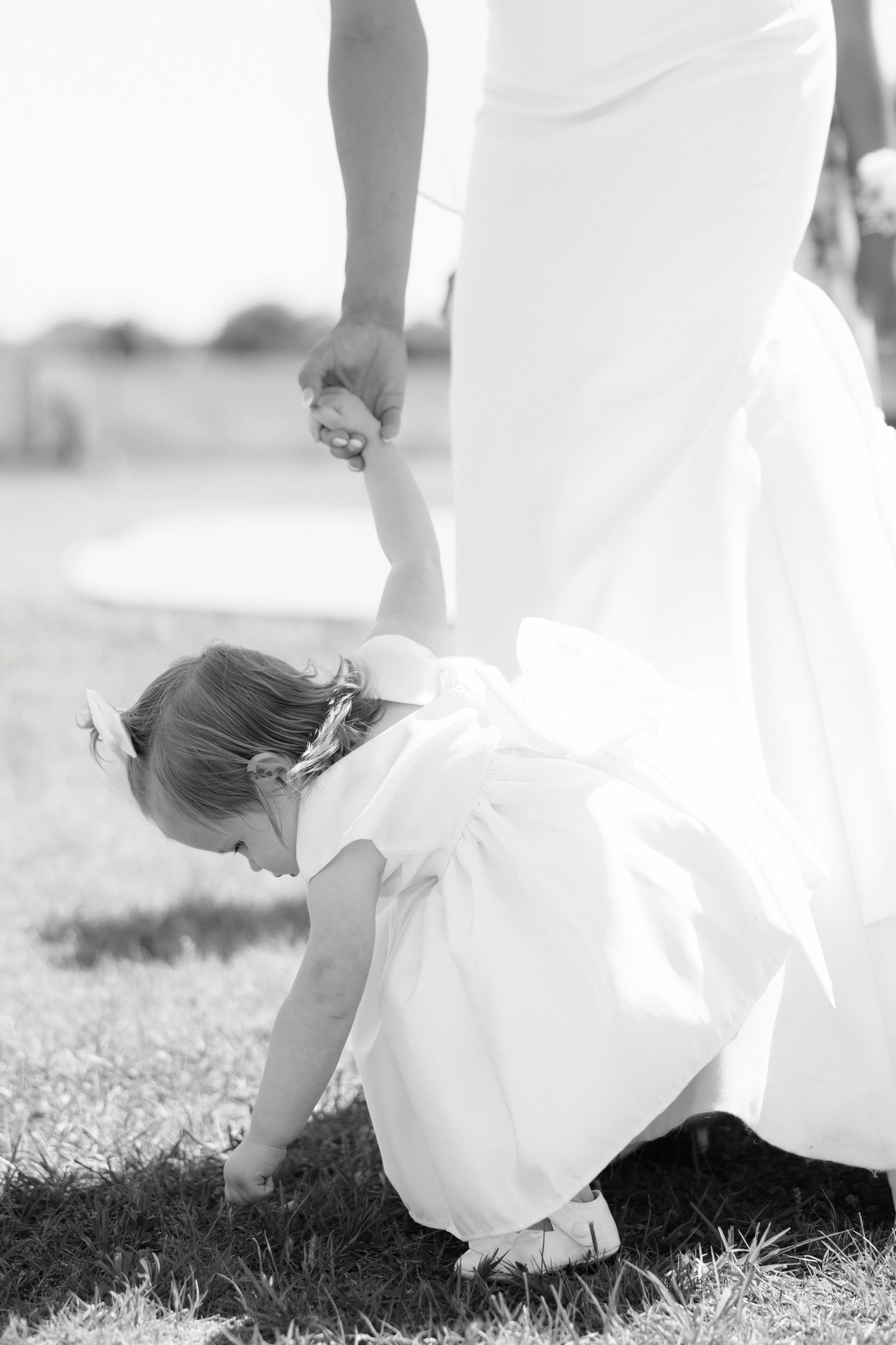 Black and white photo of flower girl bending to pick flowers while holding the bride s hand outdoors.