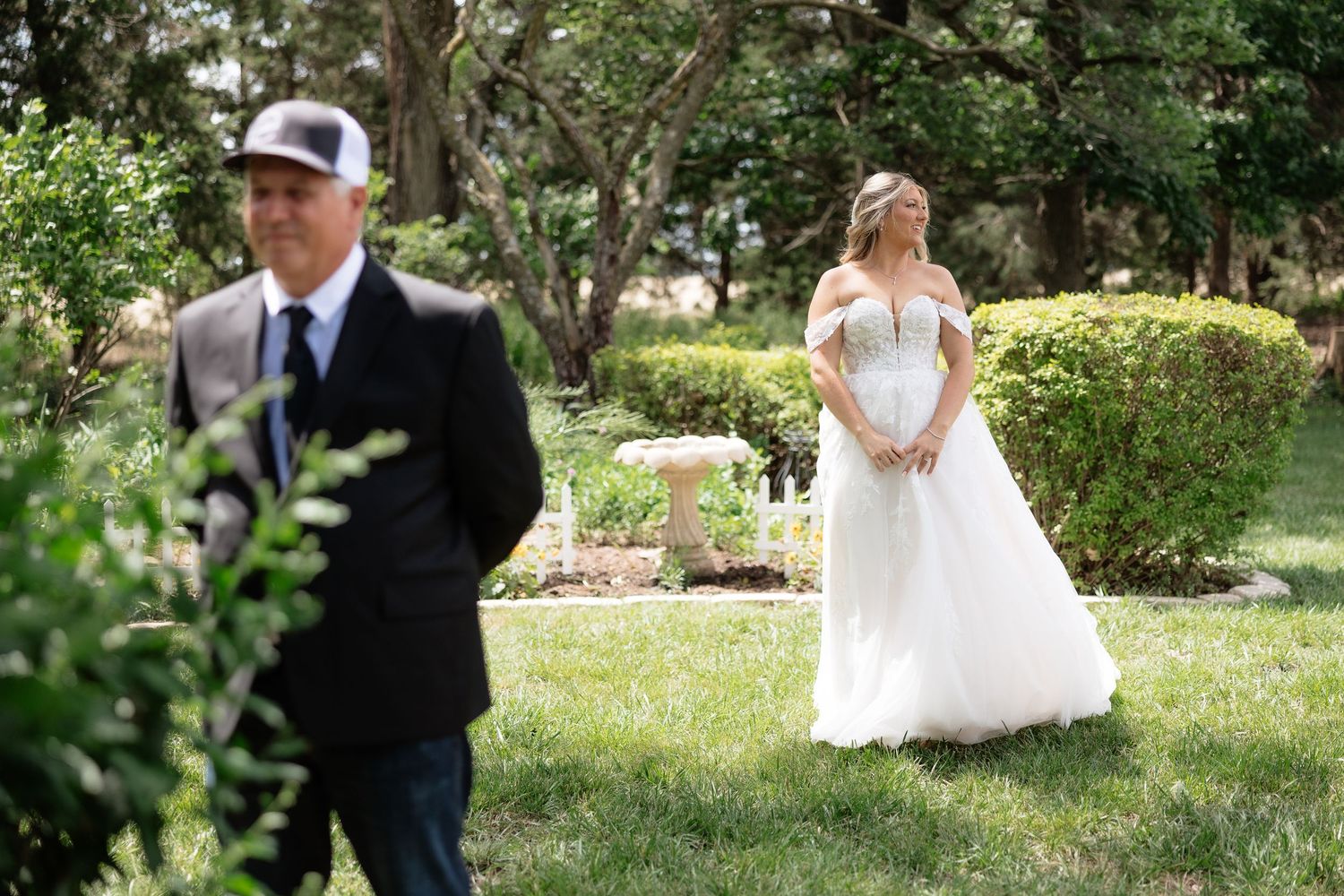 Bride in white lace gown stands behind man in black suit and cap during outdoor first look wedding moment.