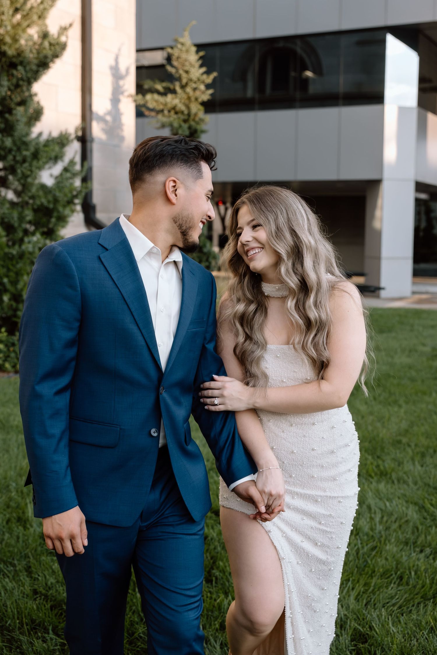 Elegant couple in formal wear smiling at each other on a green lawn.