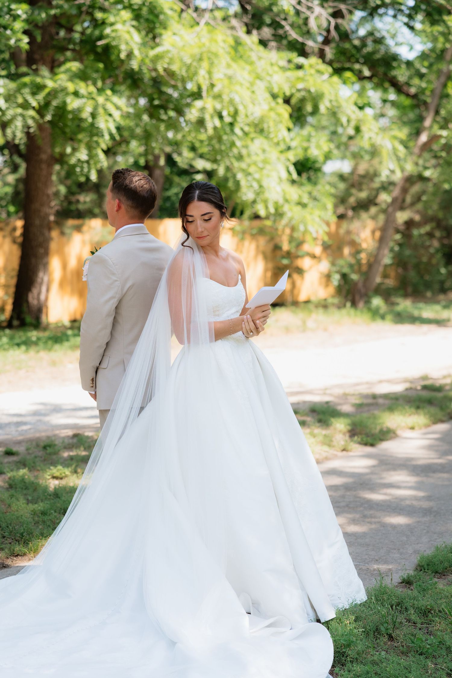 Bride in a flowing white gown reads her vows while standing behind the groom outdoors before the ceremony.