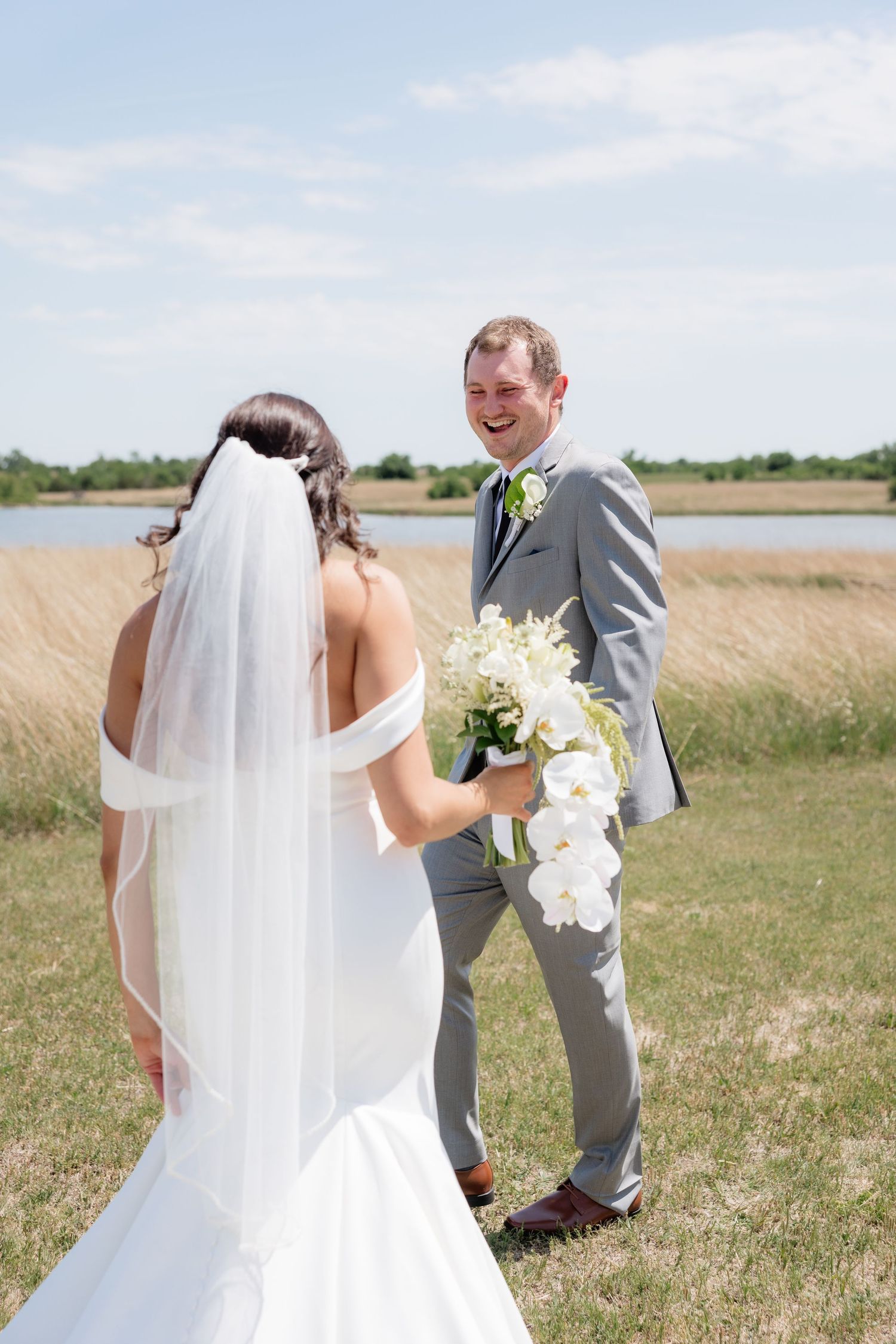 Laughing groom in gray suit reacts joyfully seeing bride in white gown during first look outdoors.