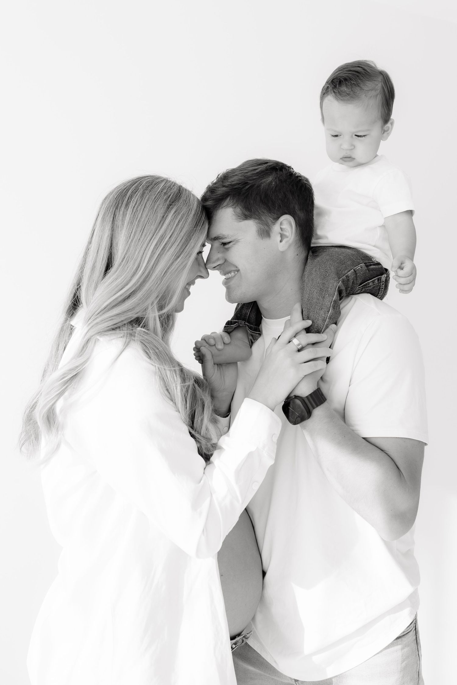 Black and white photo of a couple smiling with a toddler on the mans shoulders in a bright studio.