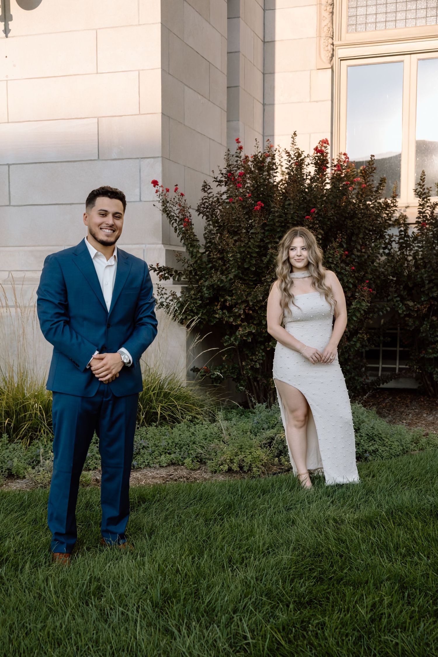 Couple posing apart on green lawn, man in navy suit and woman in white sequin dress before stone building.