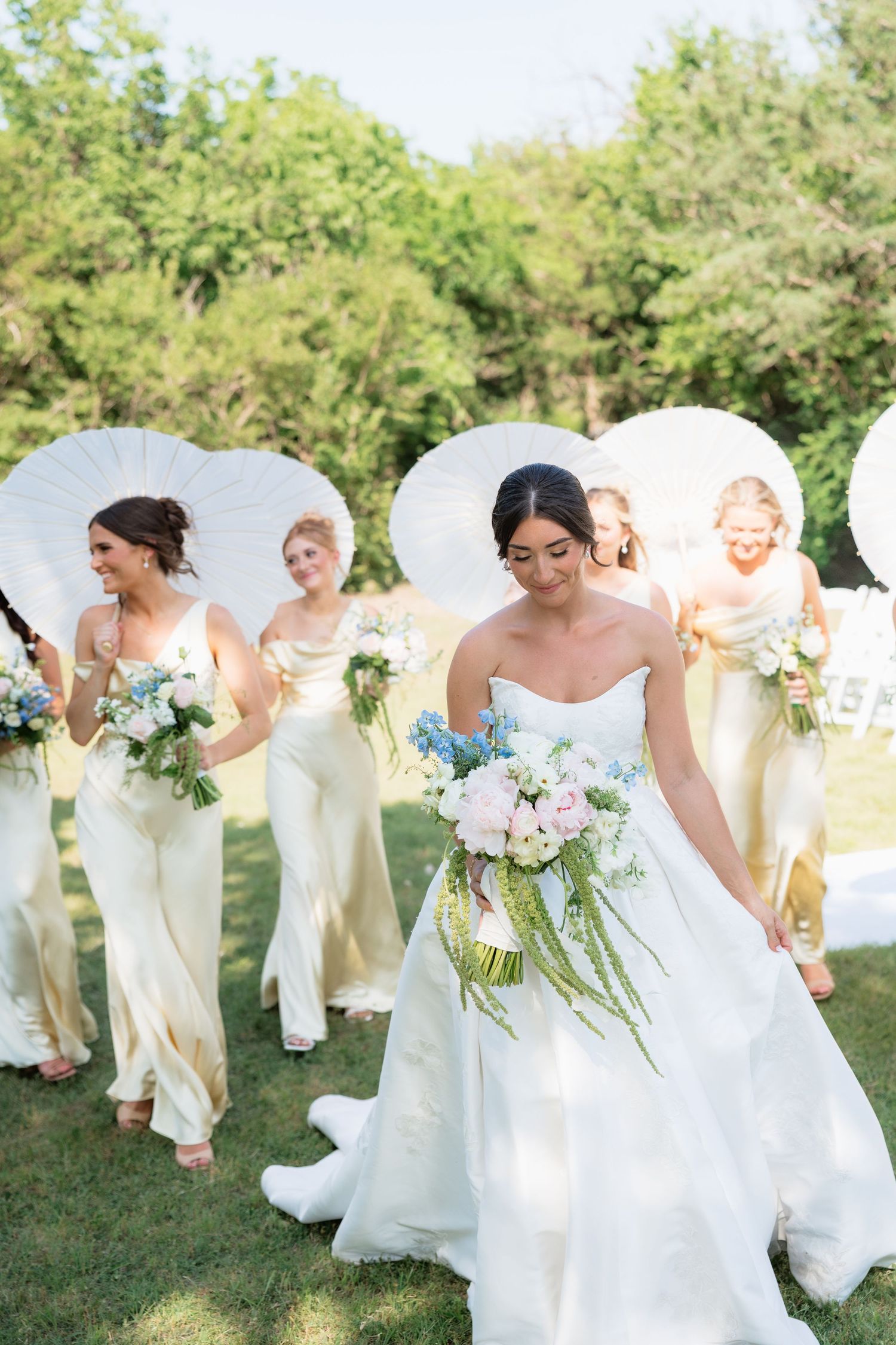 Bride in strapless gown leads bridesmaids in champagne dresses carrying bouquets and white parasols.