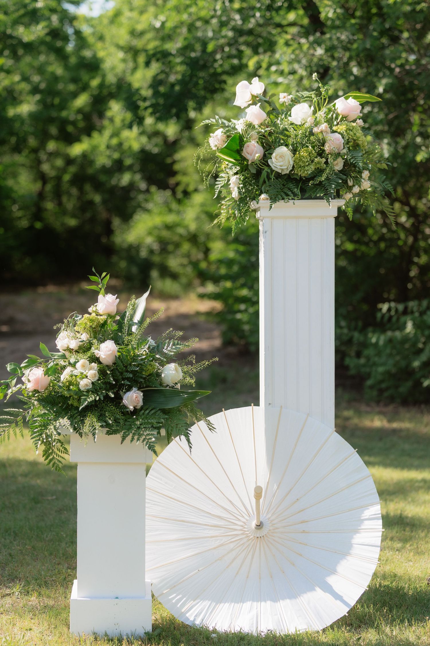 Elegant white wedding pedestals topped with lush pink rose bouquets beside a white parasol outdoors.