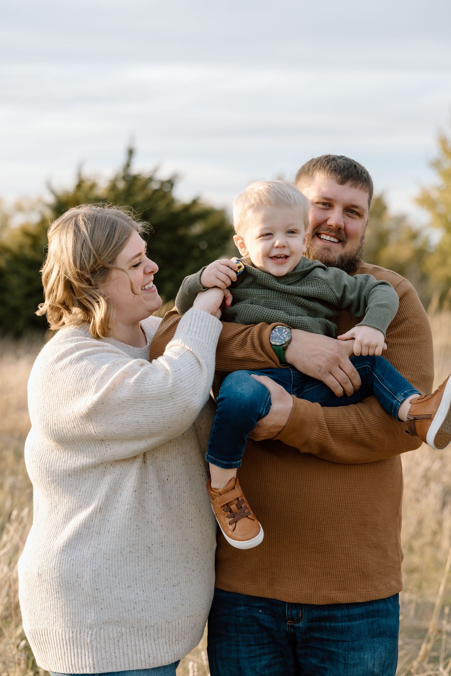 Happy young family with smiling toddler boy poses together in golden autumn field outdoors.