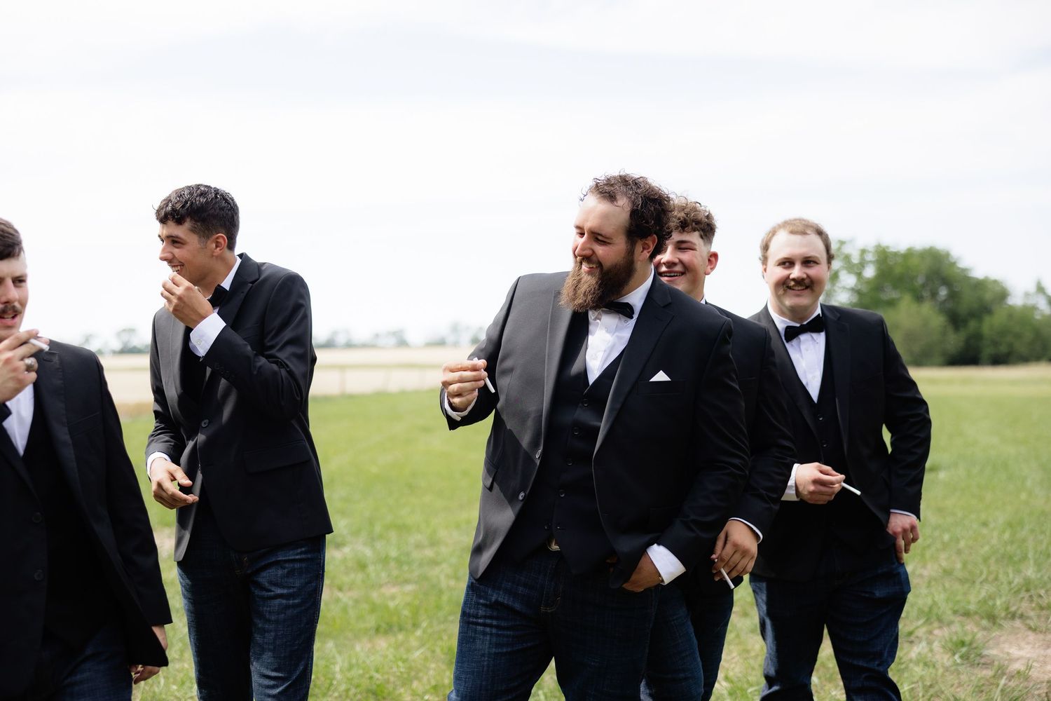 Groom and groomsmen in black tuxedos laugh and walk together across a green field on the wedding day.