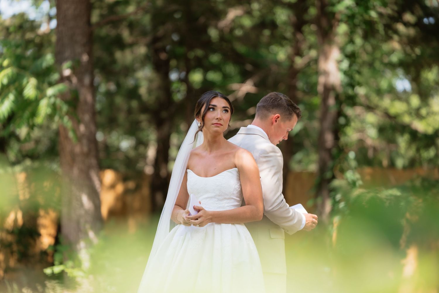 Bride in strapless white gown with veil stands in sunlit forest while groom in beige suit adjusts her dress.