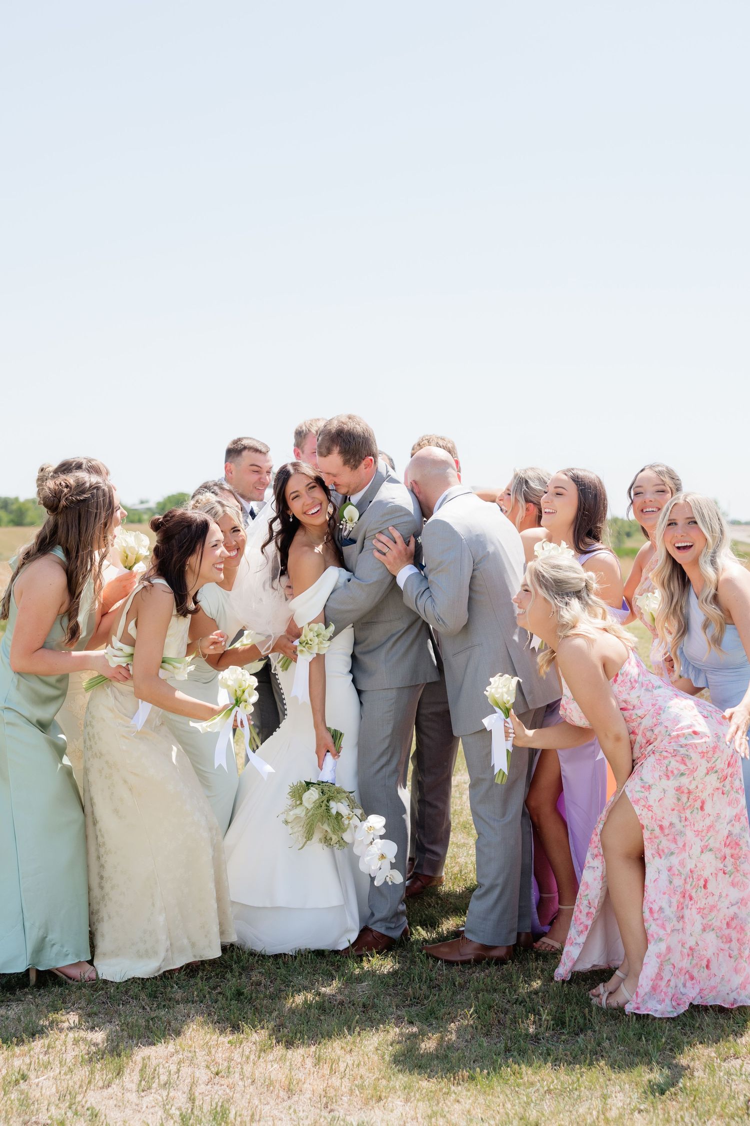 Bride and groom share a joyful kiss surrounded by their smiling wedding party in an outdoor sunny setting.