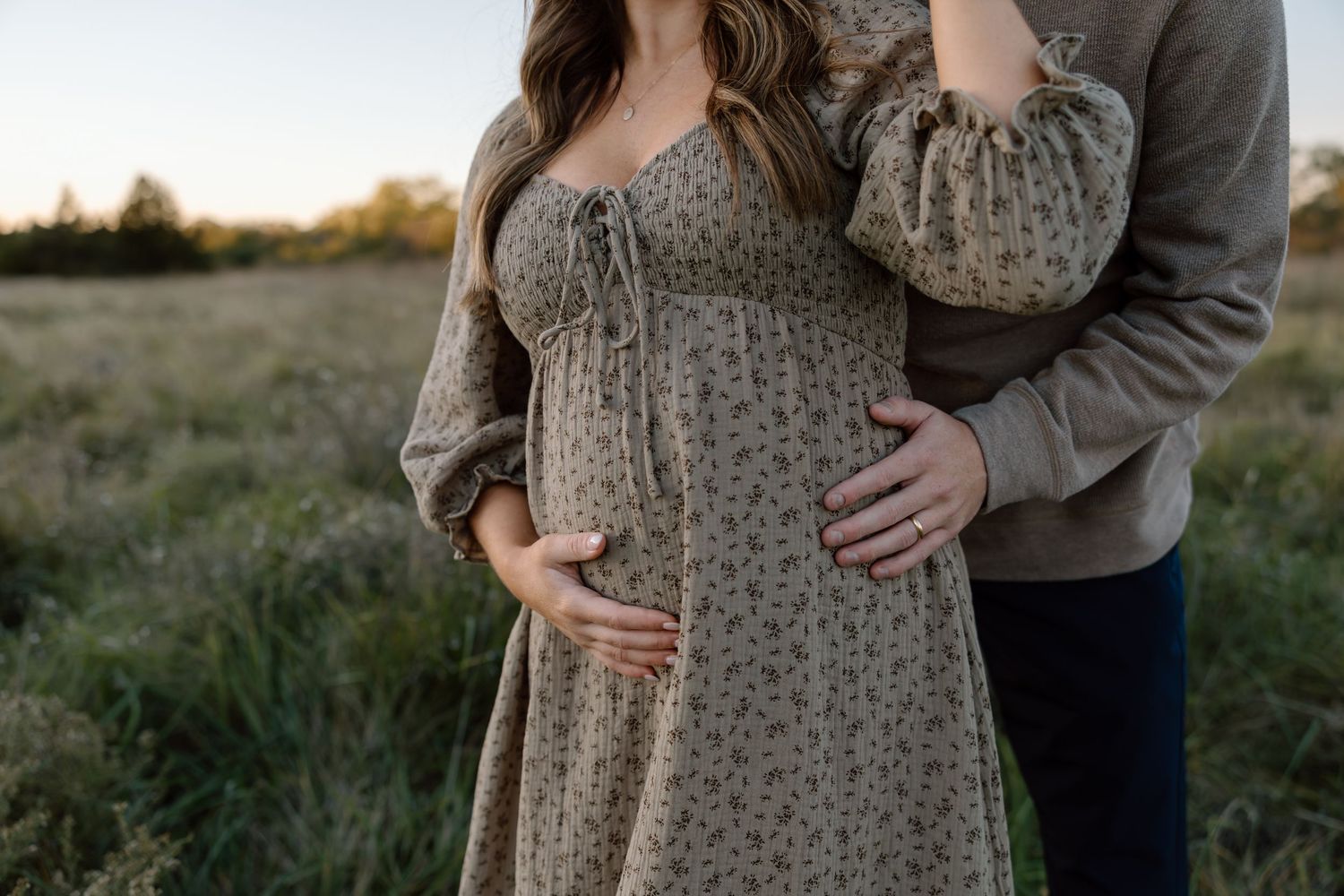 Pregnant couple standing in golden field at sunset, hands cradling baby bump tenderly.