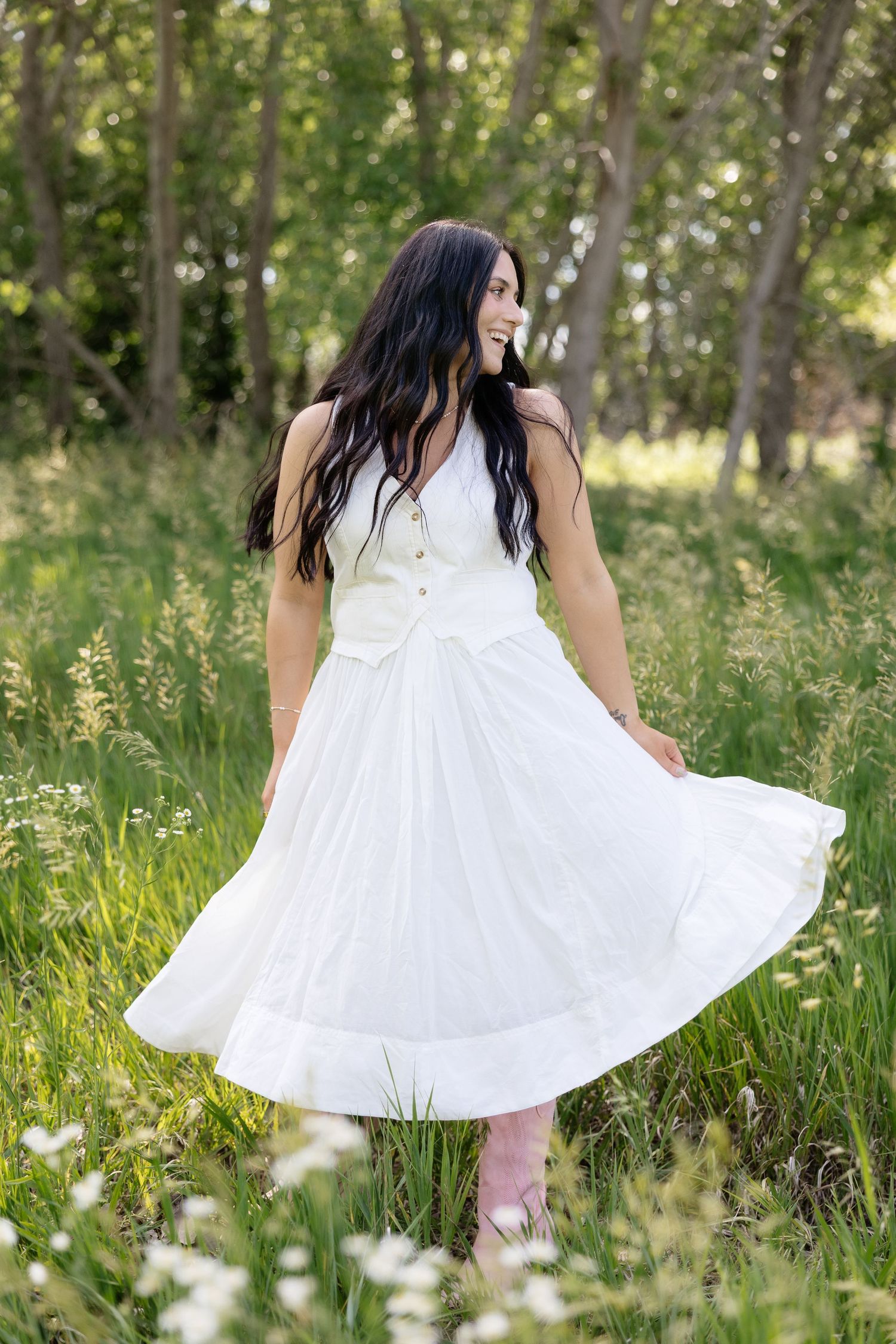 Brunette woman in white dress twirling in green meadow with wildflowers.