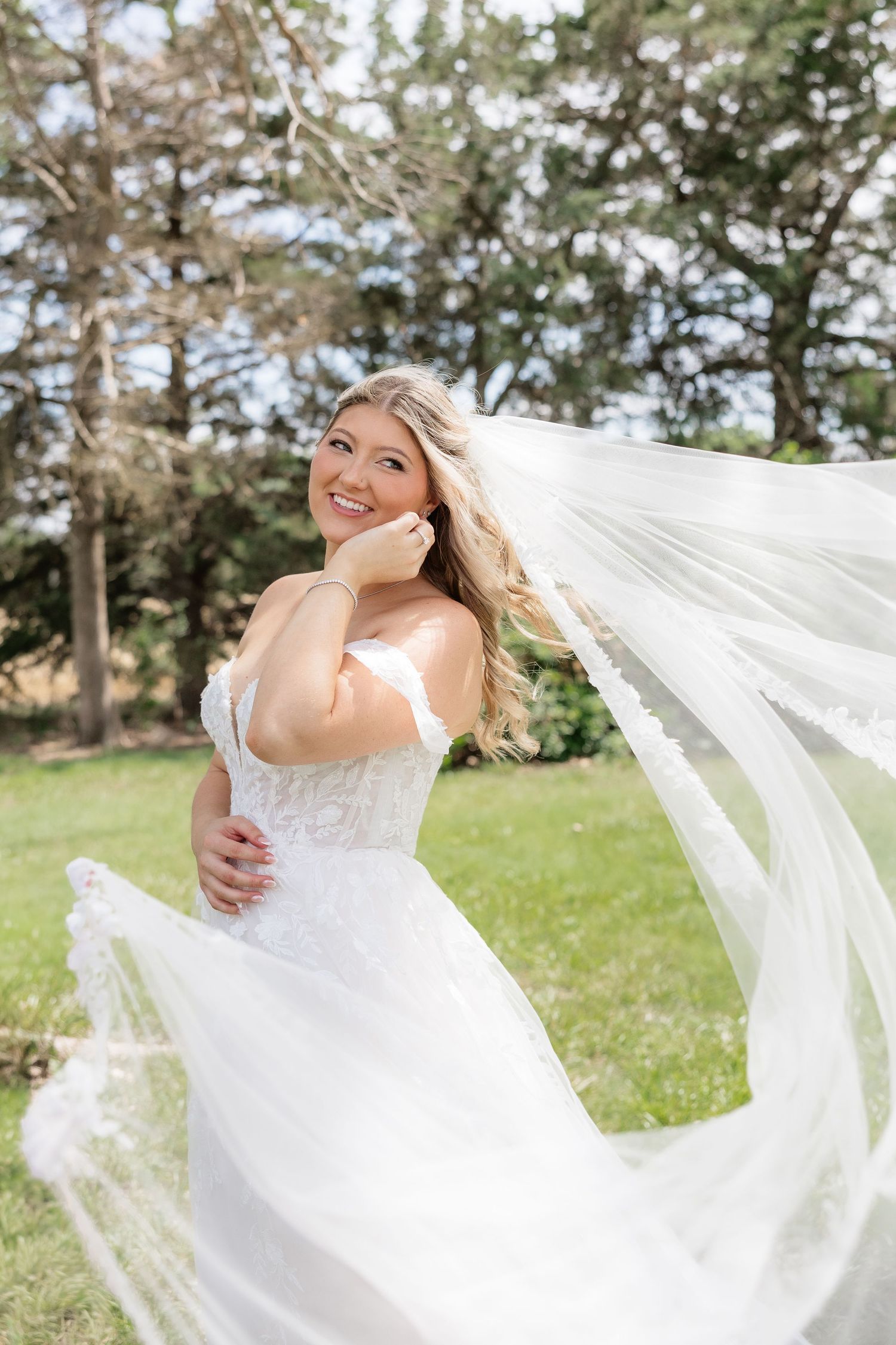 Radiant bride in off-shoulder lace gown smiles as her veil flows in the breeze in a sunlit garden.