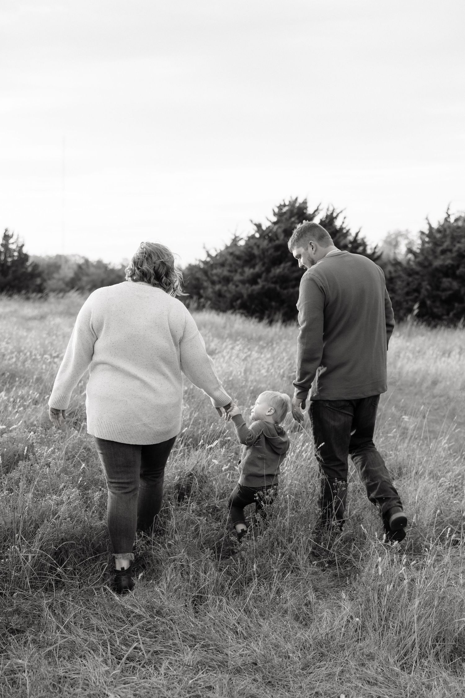 Black and white photo of parents holding hands with their toddler child while walking through an autumn field.