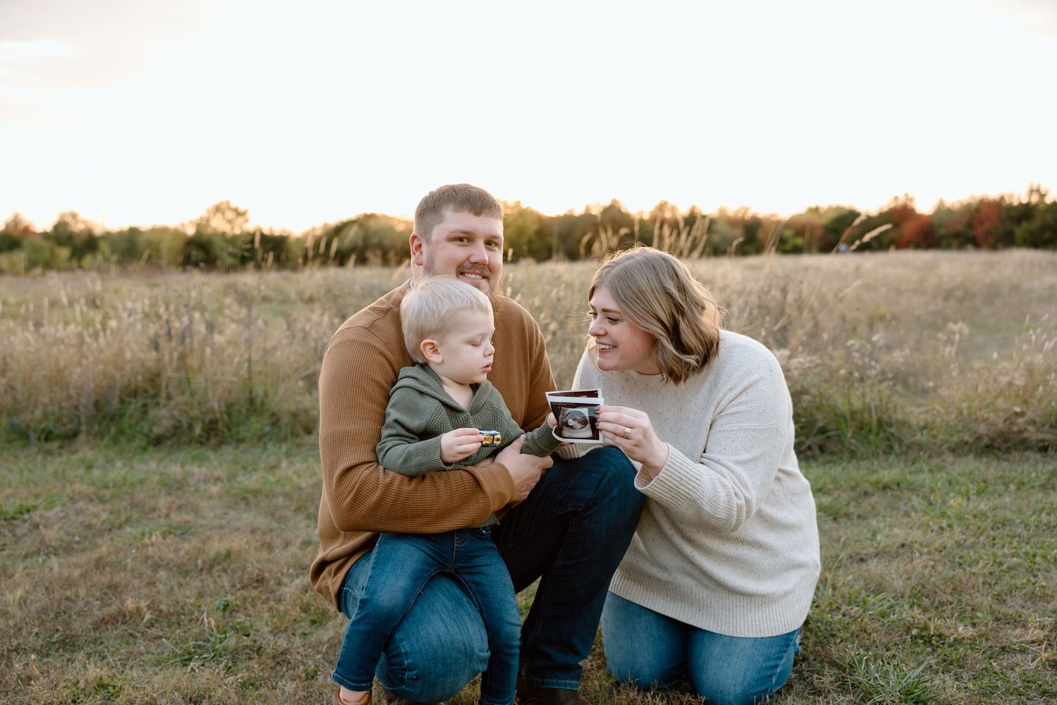 A happy family of three sits in a golden field at sunset, parents showing ultrasound photos to their toddler son.