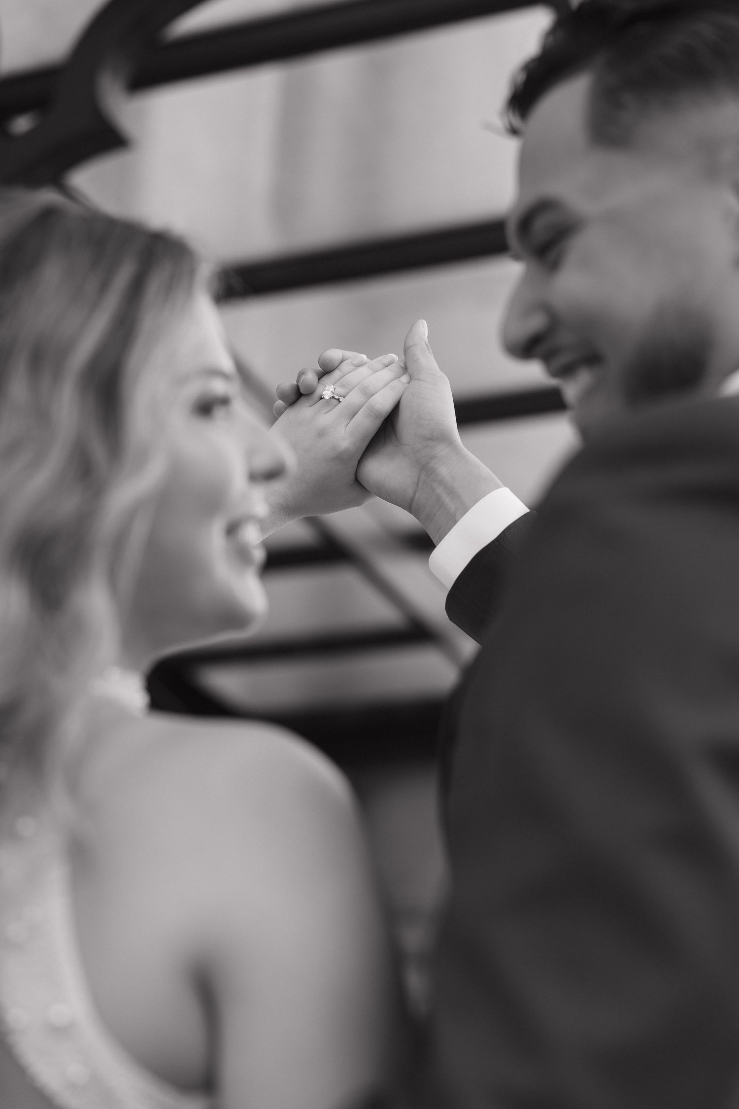 Black and white photo of smiling couple holding hands showing engagement ring.