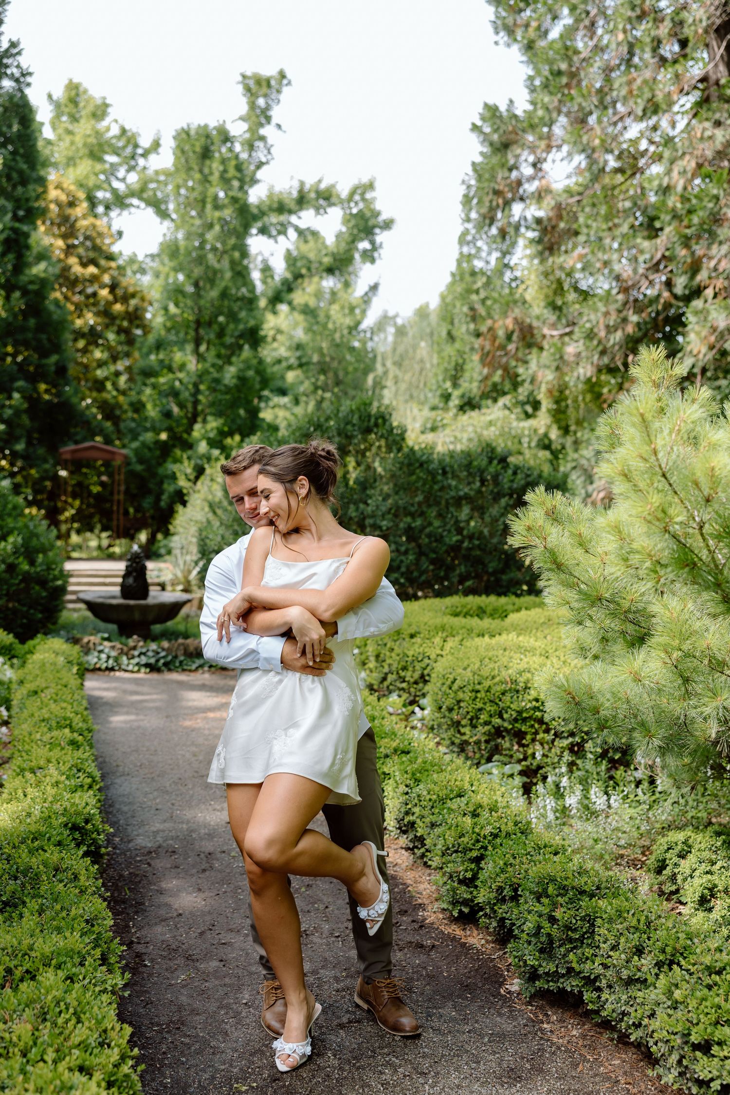 Couple embracing on a garden path surrounded by lush green shrubs and trees.