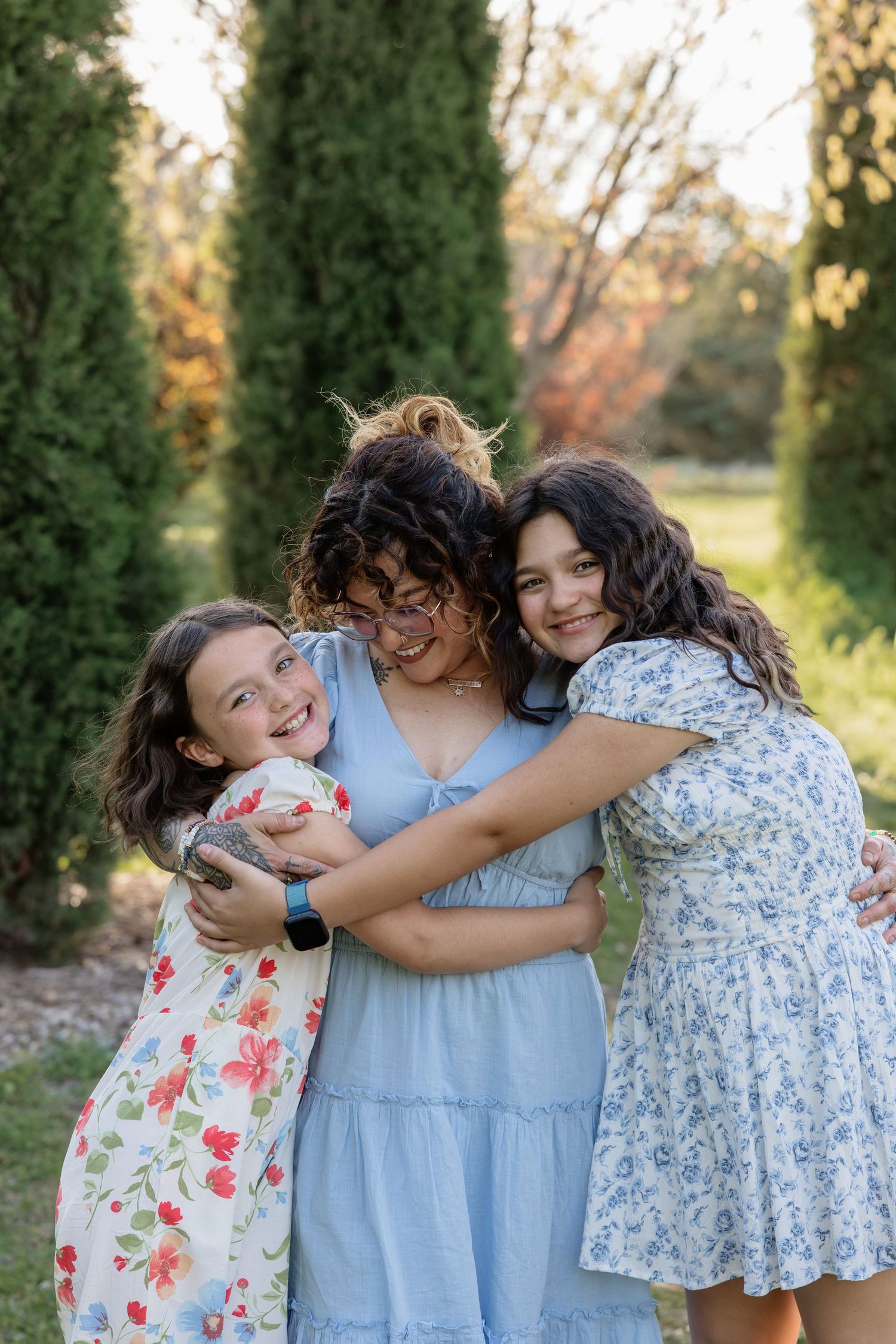 A joyful woman in a blue dress receives warm hugs from two smiling girls in a lush green garden.