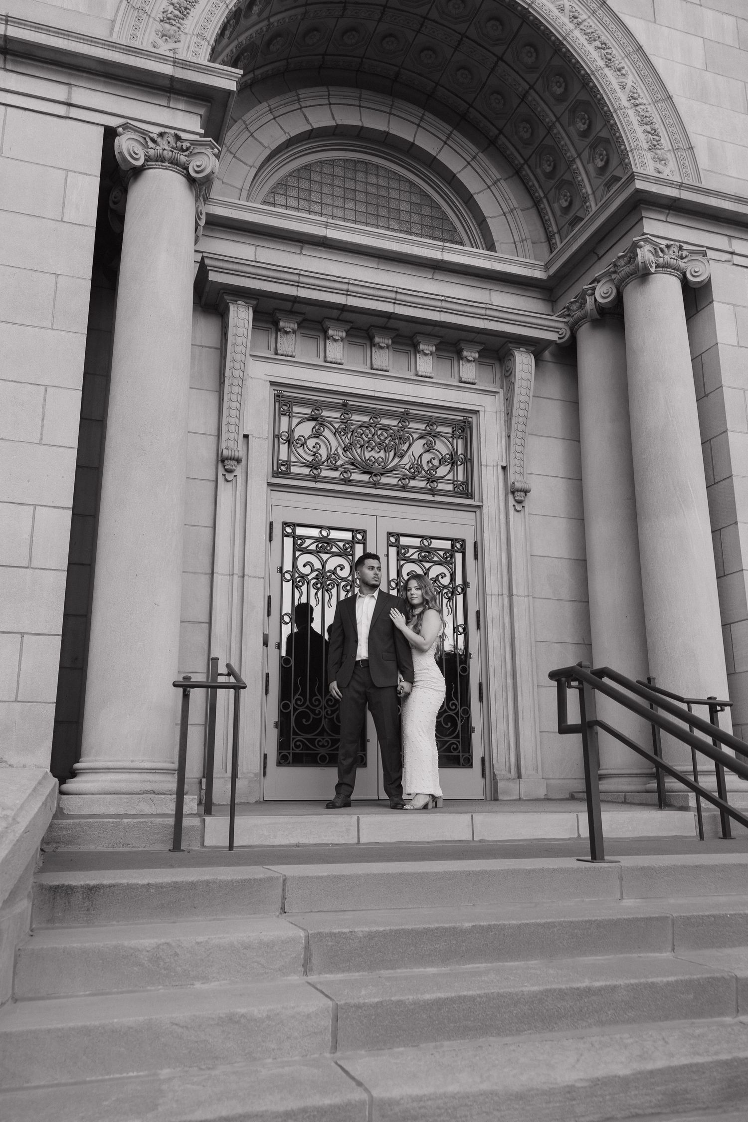 Black and white photo of elegant couple posing on steps of grand building entrance.