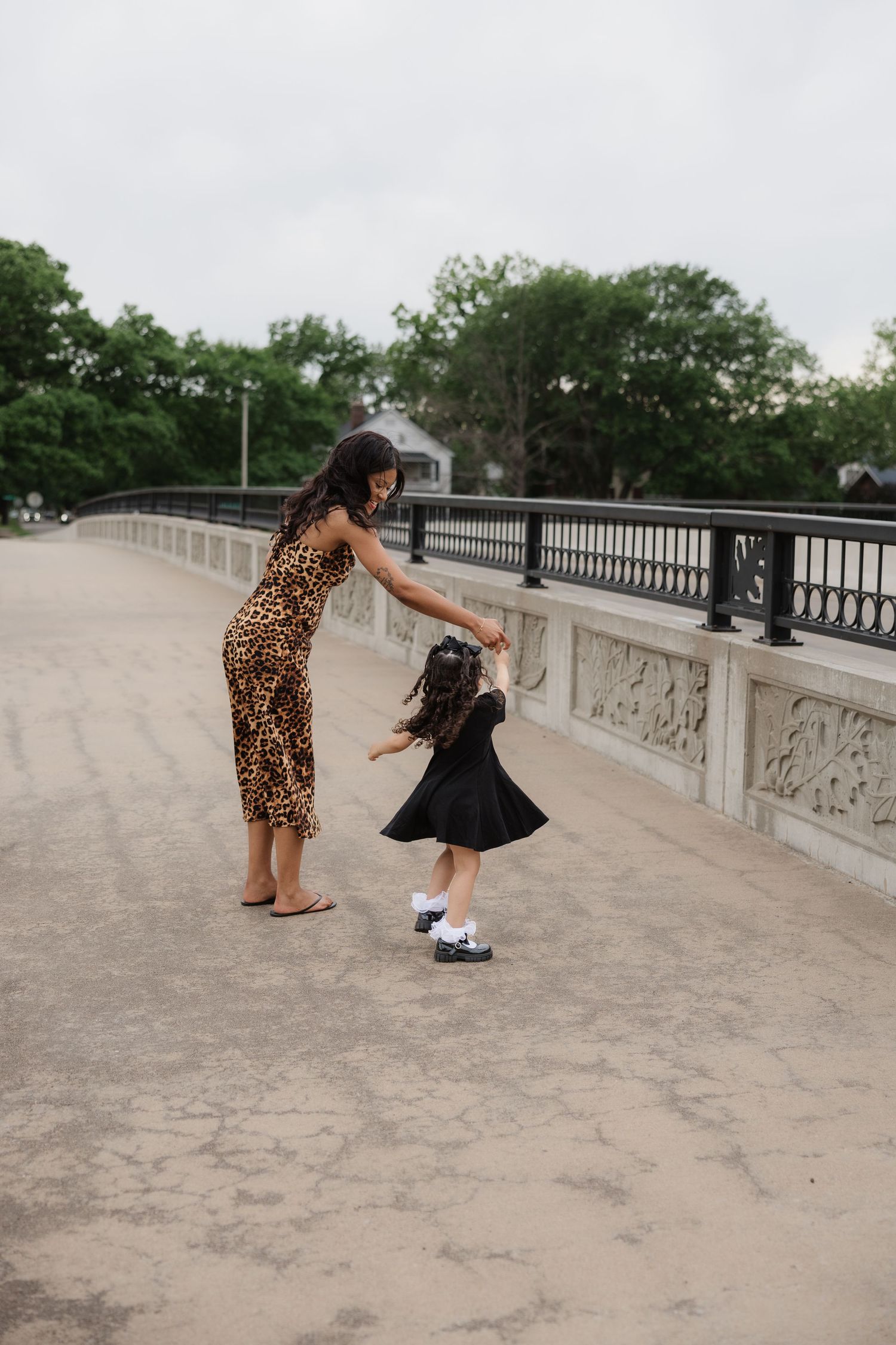 A woman in leopard print dress dances with a toddler in a black dress on a decorative bridge outdoors.