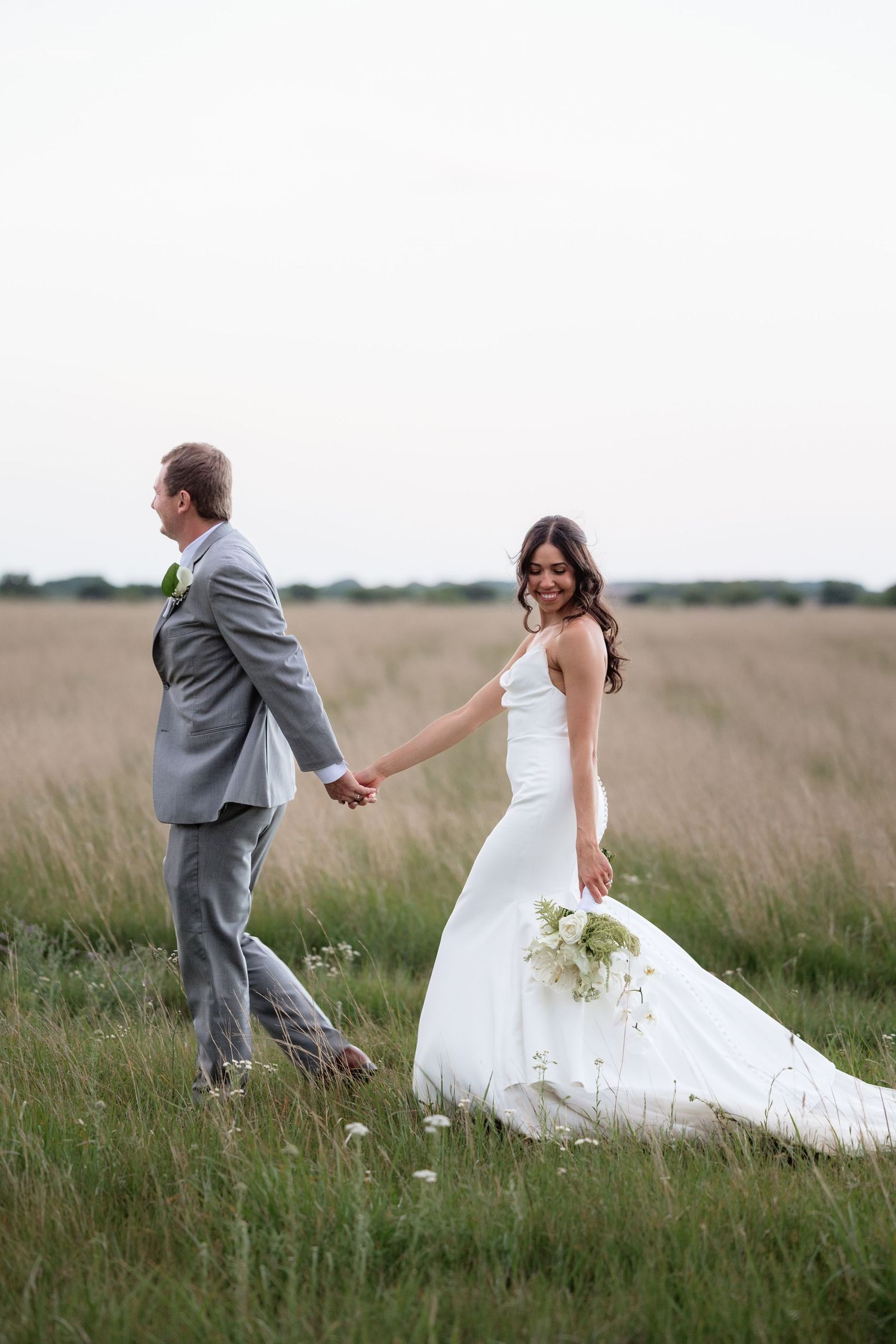 Bride and groom walk hand in hand through golden meadow on wedding day.