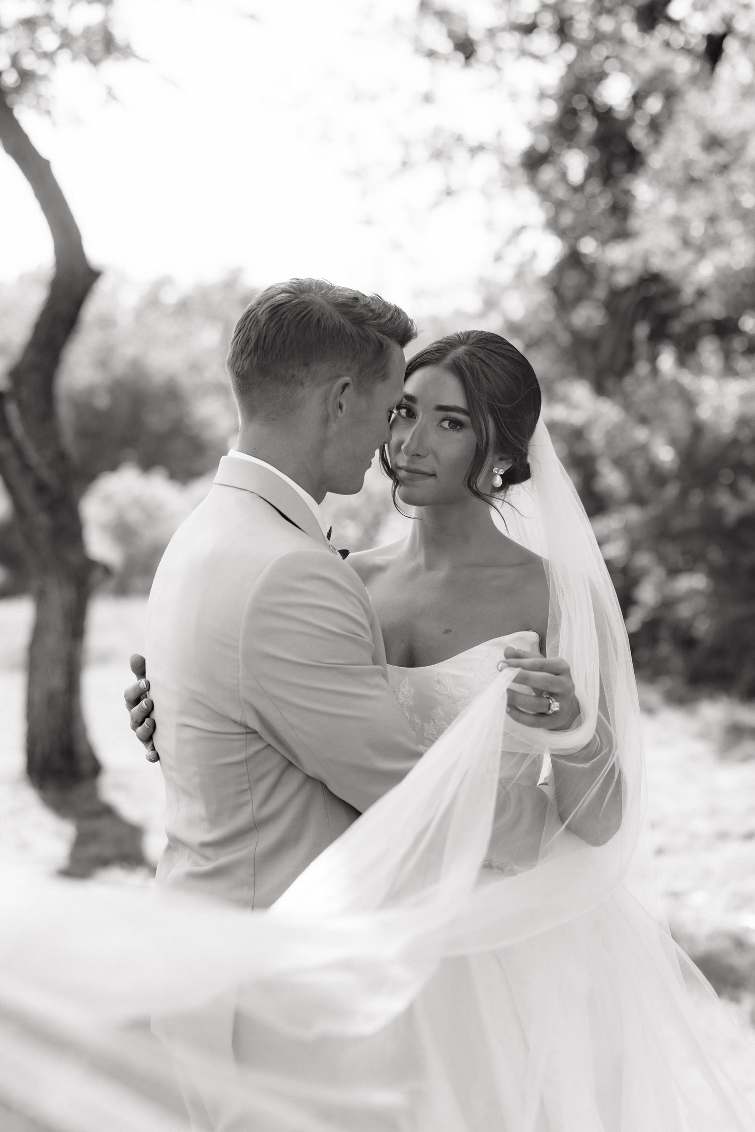 Black and white photo of bride and groom embracing outdoors, veil flowing in the breeze among trees.