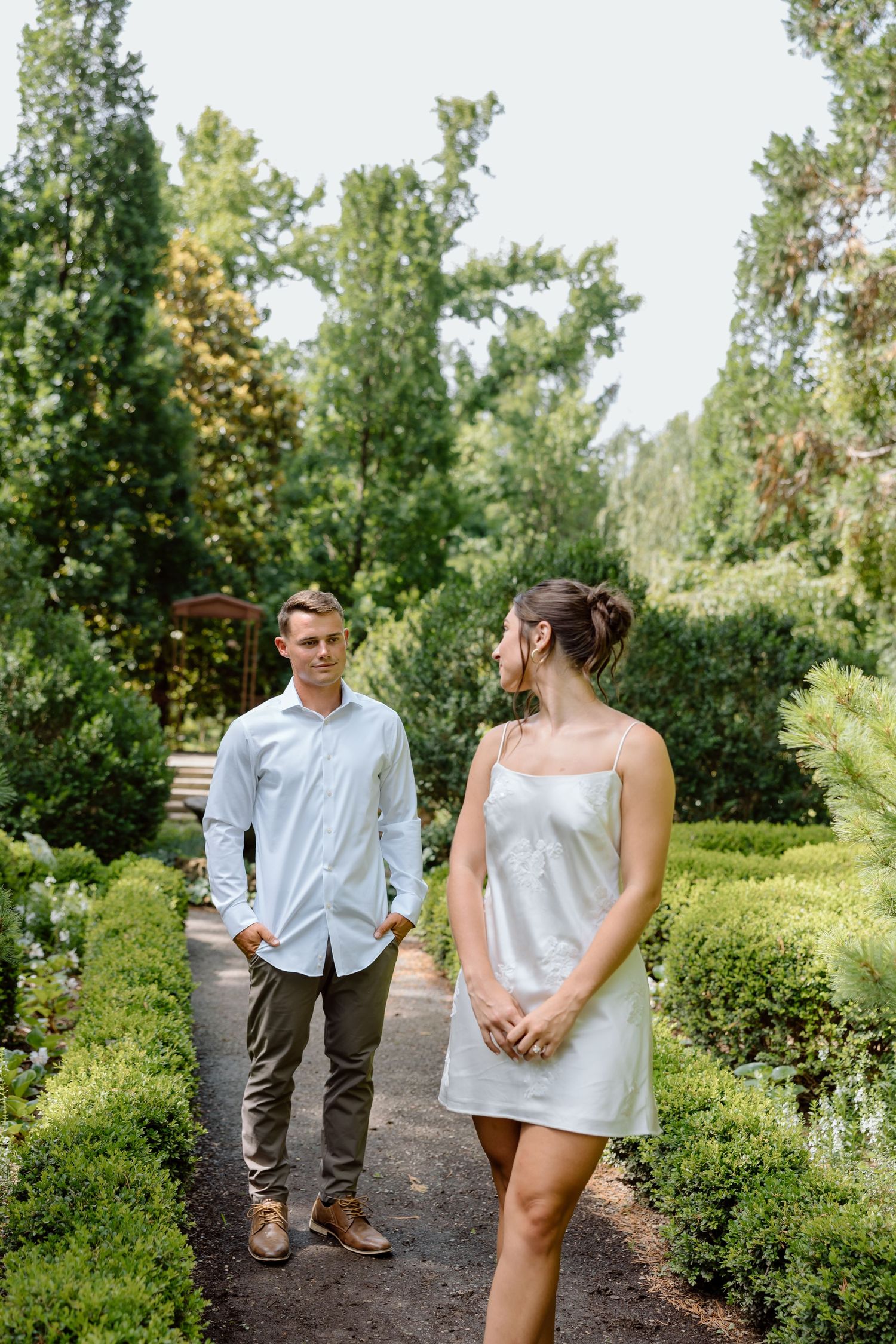 Couple standing apart on a garden path surrounded by lush green trees and manicured hedges.