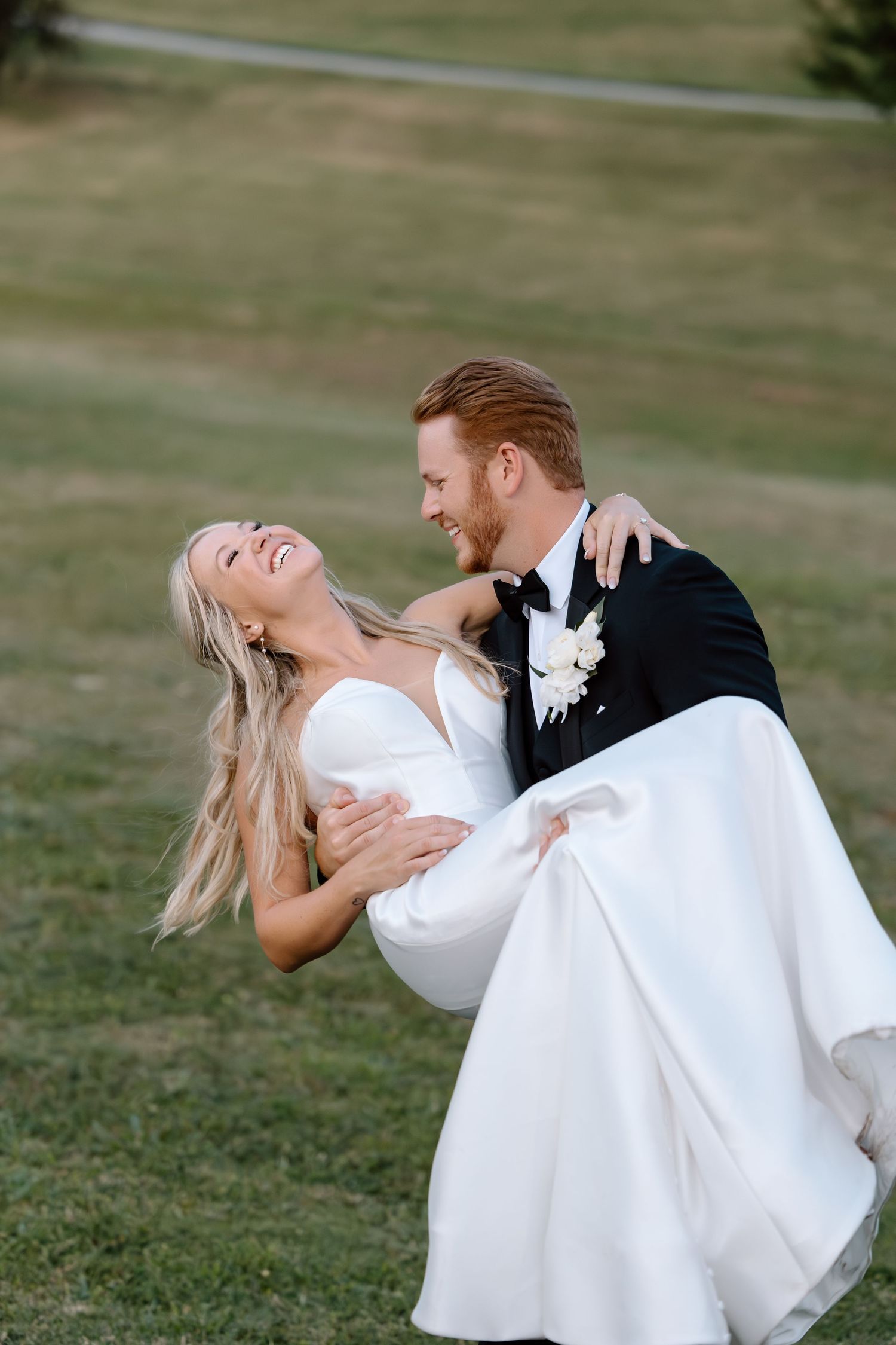 Groom lifting laughing bride in white wedding gown on green lawn during outdoor celebration.
