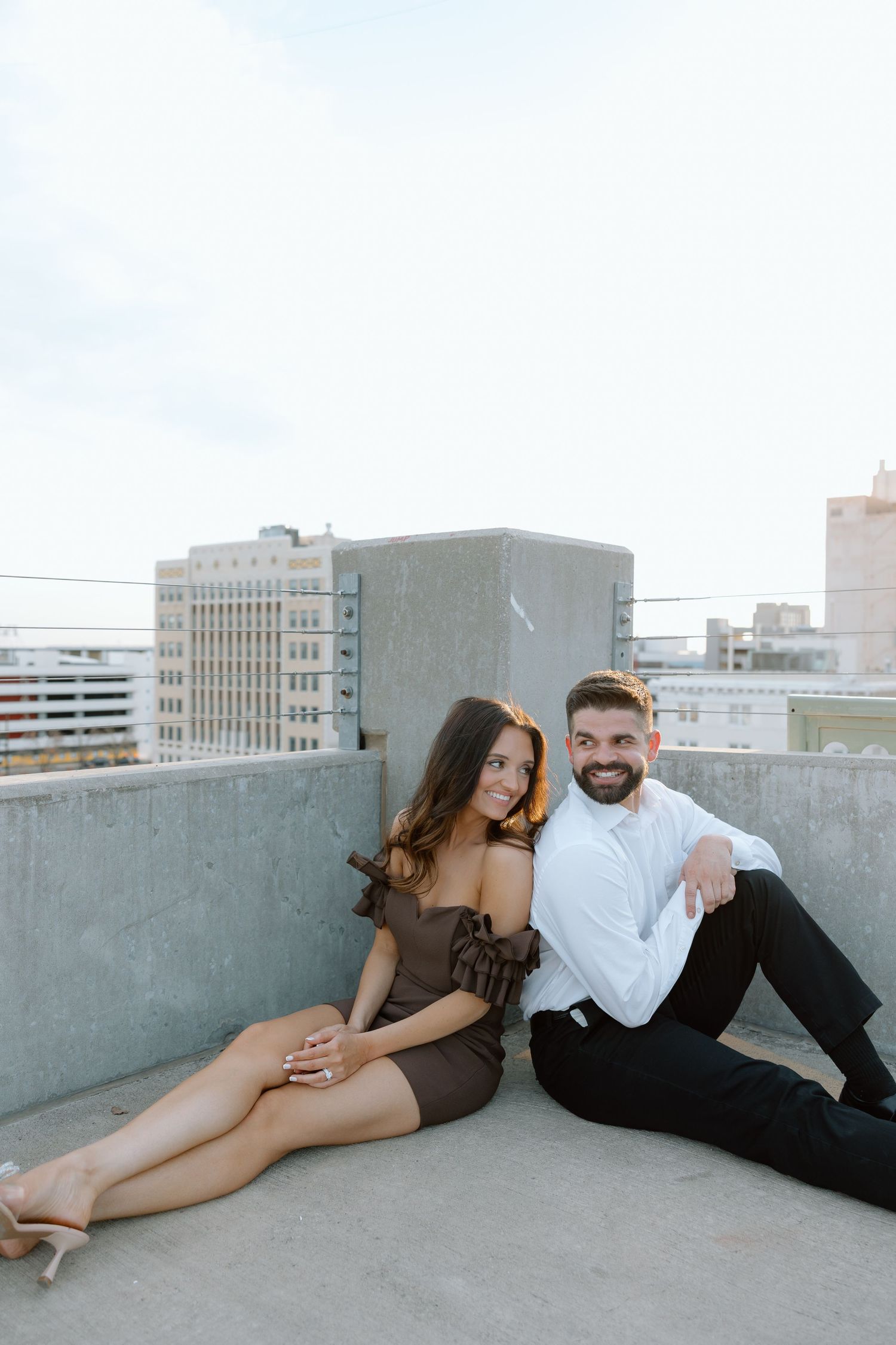 Smiling couple posing together on rooftop with city buildings, woman in brown off-shoulder dress.
