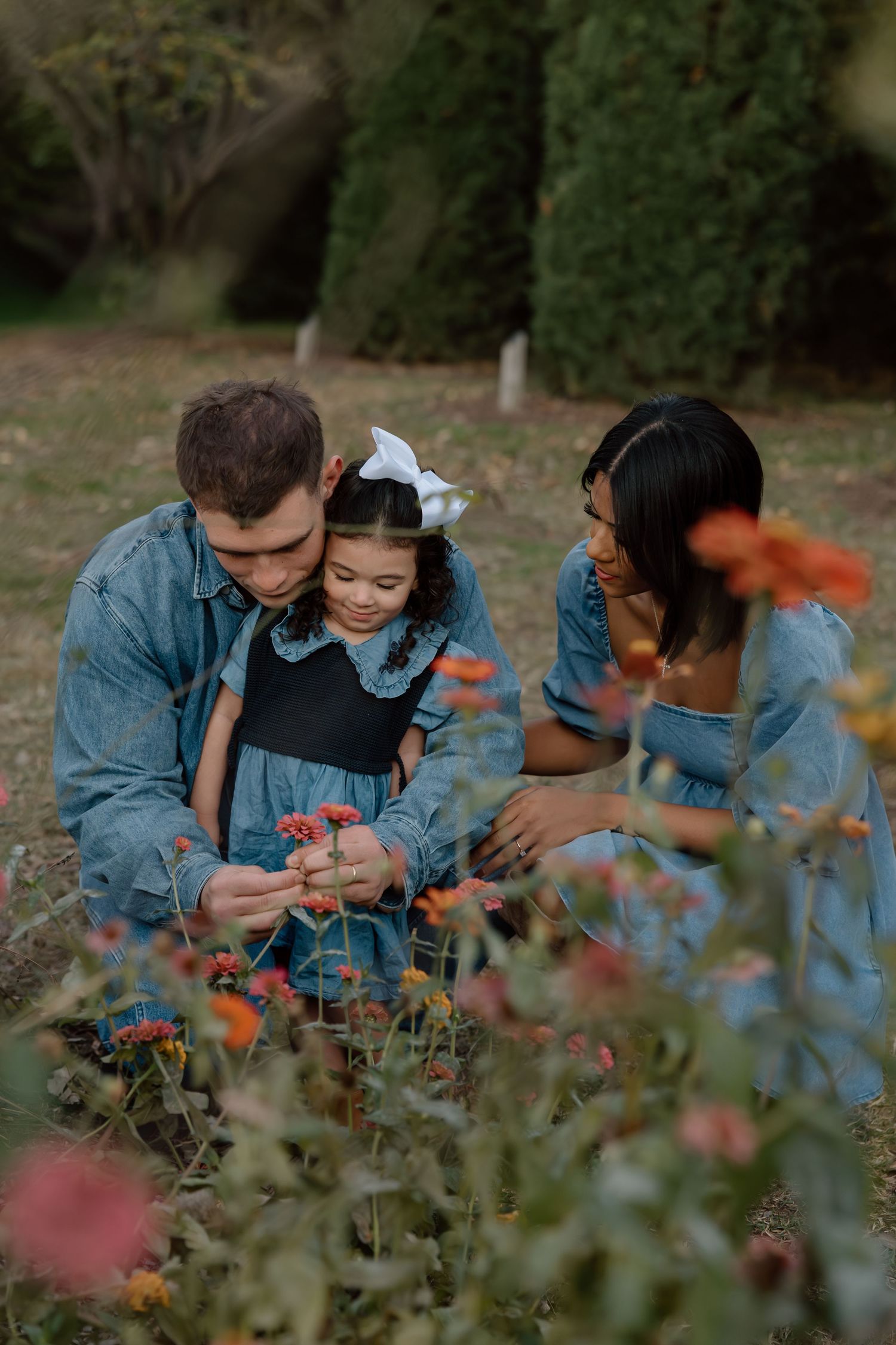 A family in denim outfits crouches together to examine colorful flowers blooming in a garden.
