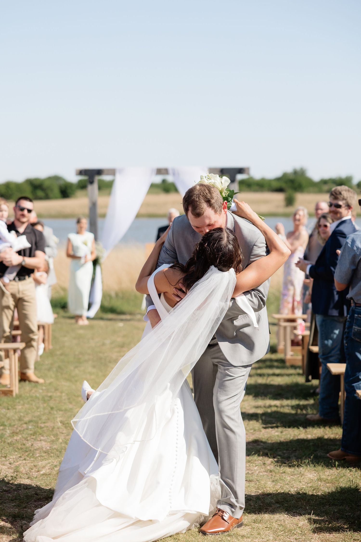 Groom dips bride for romantic kiss after outdoor wedding ceremony by lake.