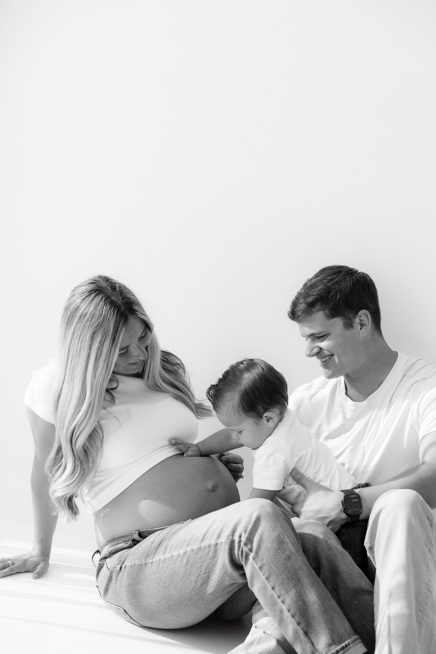 Black and white image of a pregnant woman and man sitting as a toddler kisses her bare baby bump.