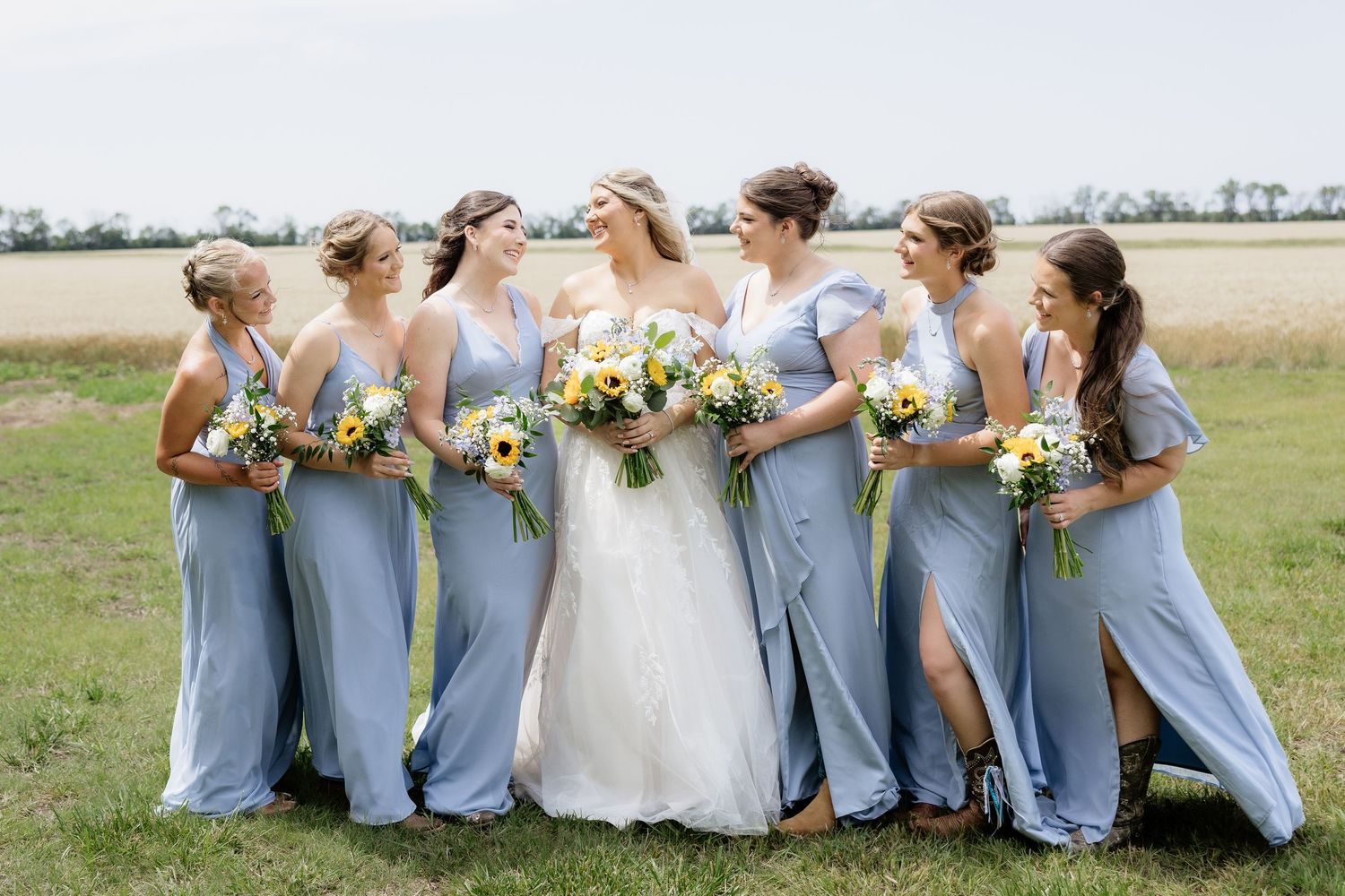 Bride with bridesmaids in dusty blue gowns holding yellow and white bouquets pose together in a field.