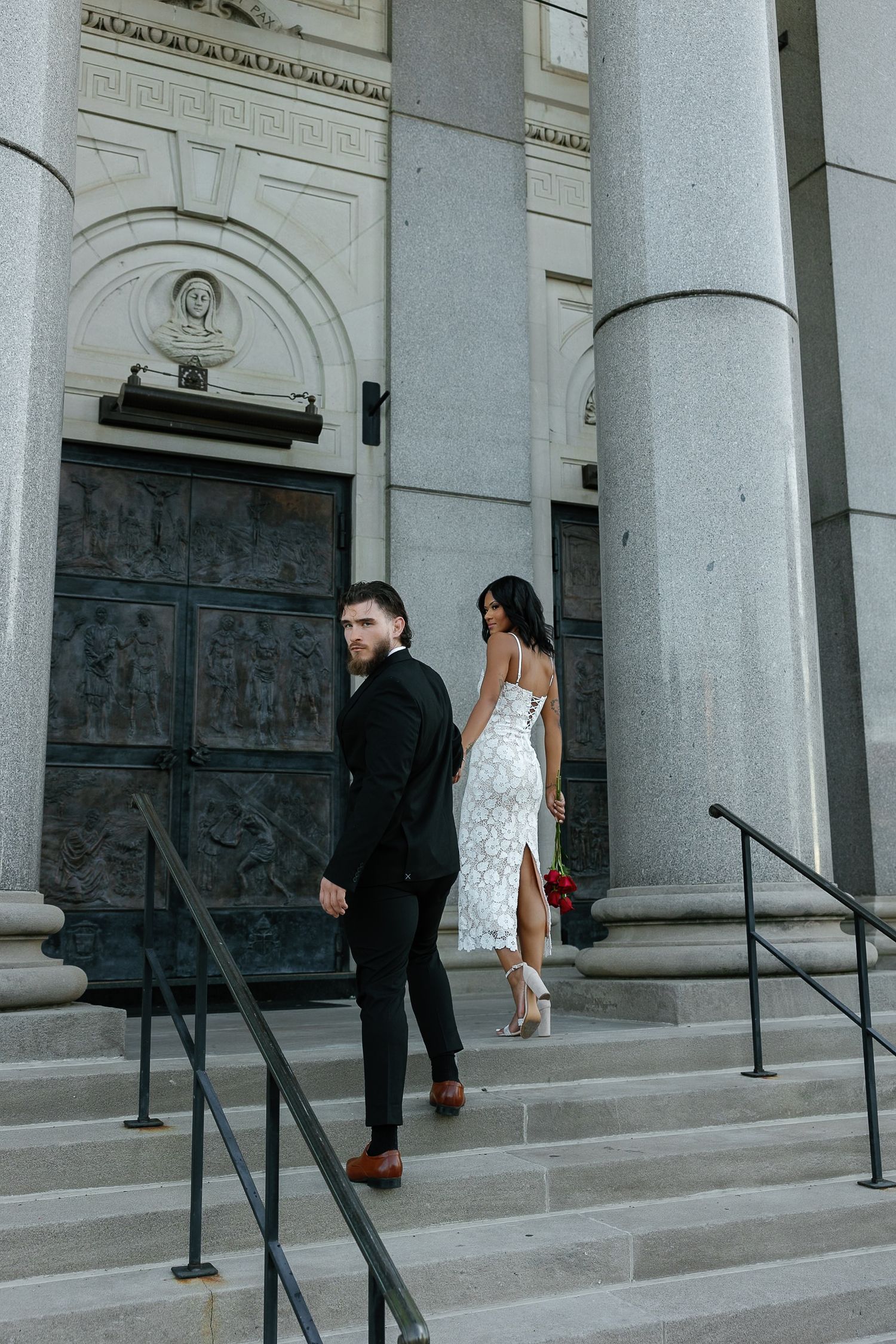 Elegant interracial couple descending church steps, woman in white lace dress holding red roses.