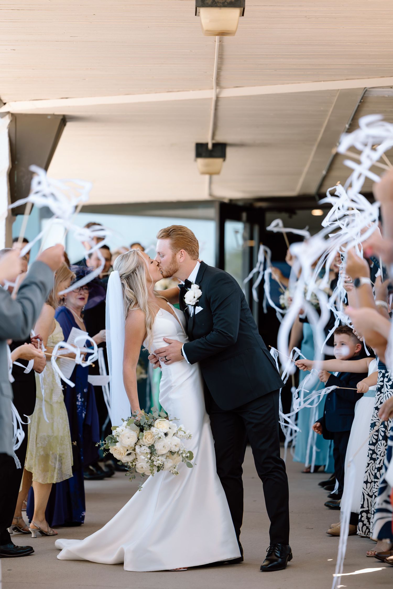 A bride and groom share a kiss during their wedding sendoff while guests wave white ribbon wands.