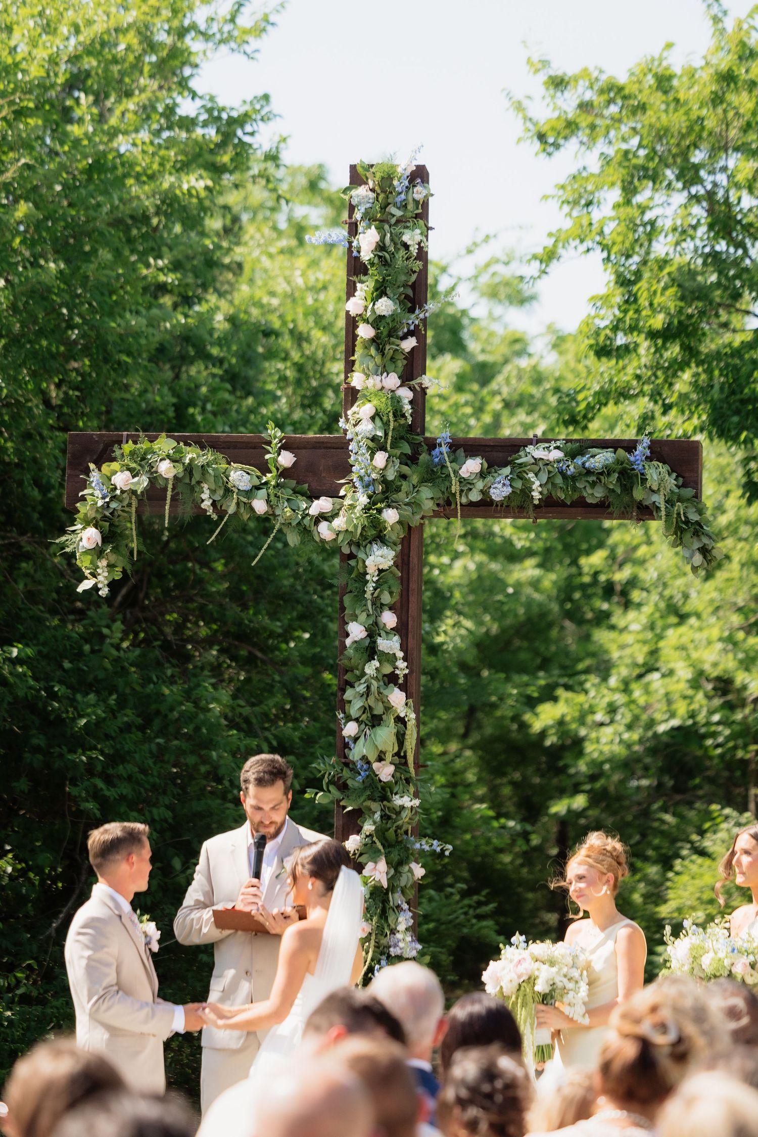 Bride and groom exchange vows at outdoor ceremony beneath a large wooden cross decorated with flowers.