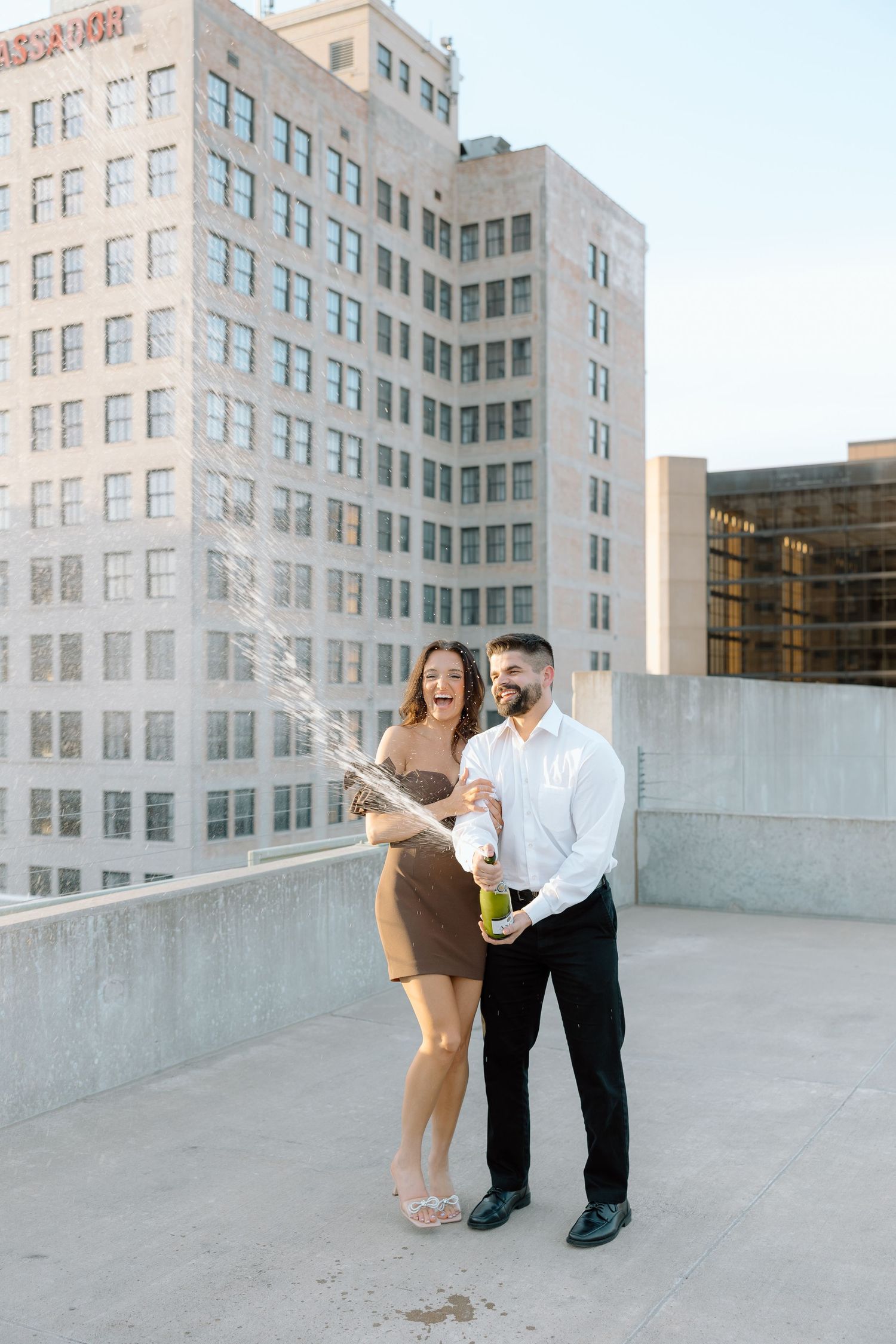 Joyful couple popping champagne bottle on rooftop with city buildings behind them.