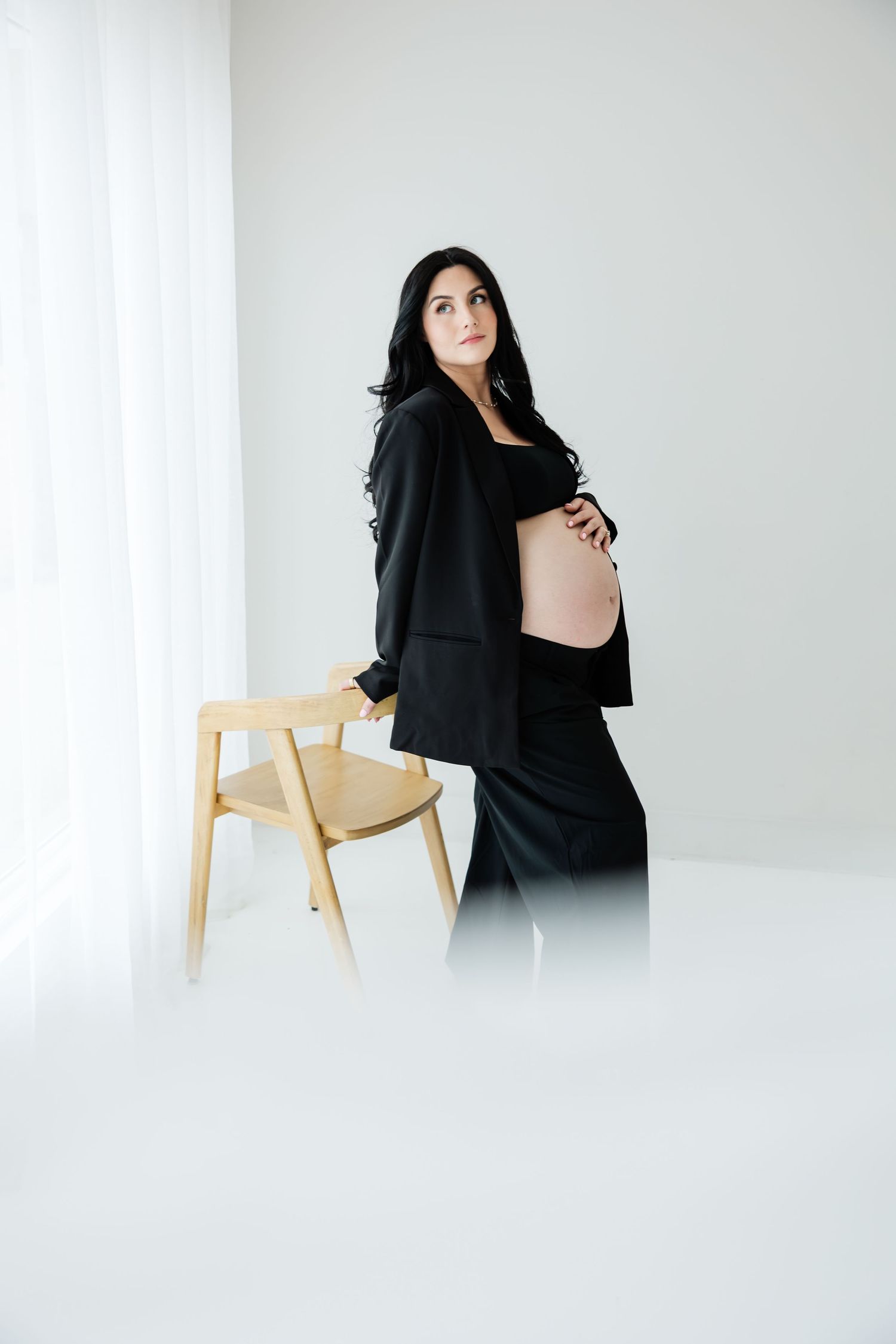 Elegant pregnant woman in black outfit cradling her baby bump beside a wooden chair in white room.