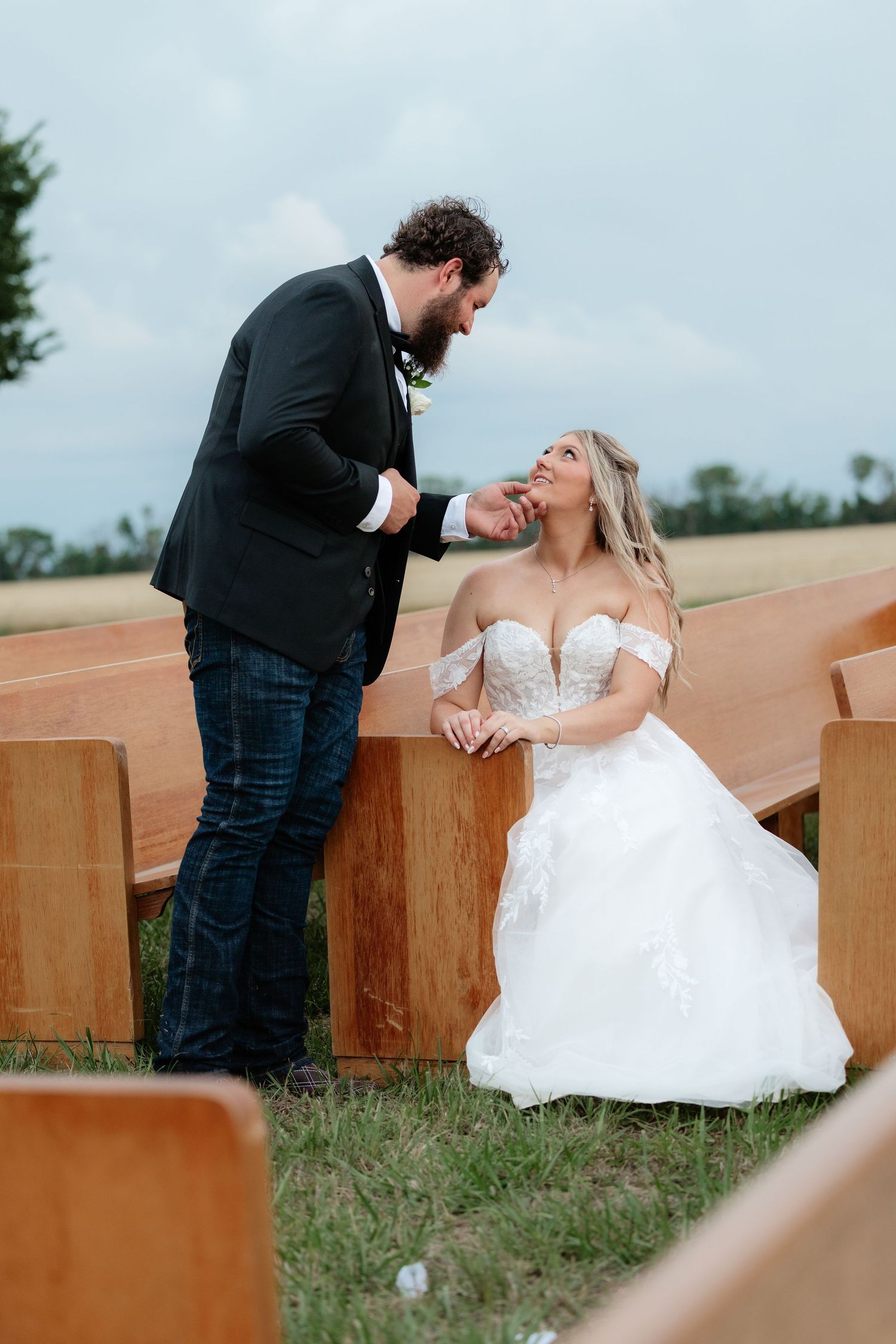 Bearded groom leans over wooden pew smiling at seated blonde bride at outdoor wedding venue.